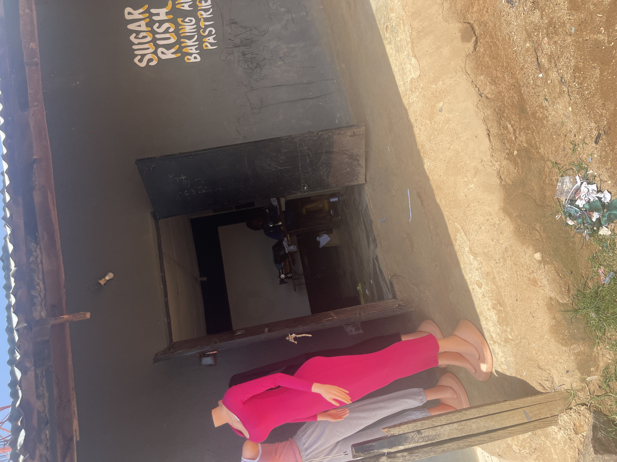 The image depicts the exterior of a small business establishment, likely a shop, in Rafin Par, Nigeria. A corrugated metal roof extends over the storefront, supported by wooden beams. The building's facade is painted dark gray. To the right of the entrance, white and yellow text is painted on the wall, reading "SUGAR RUSH BAKING AND PASTRIES".
In front of the shop, two mannequins are displayed. The mannequin on the left wears a pink and white striped tank top and gray sweatpants. The mannequin on the right is dressed in a long, form-fitting pink dress. Both mannequins are mounted on oval bases.
The entrance to the shop is open, revealing a glimpse of the interior. Inside, a person is seated at a table with a sewing machine, suggesting the business might also offer tailoring services. The floor area in front of the shop is a dirt and gravel surface, with some scattered debris, including plastic waste and greenery. The overall impression is that of a humble, functional commercial space.
