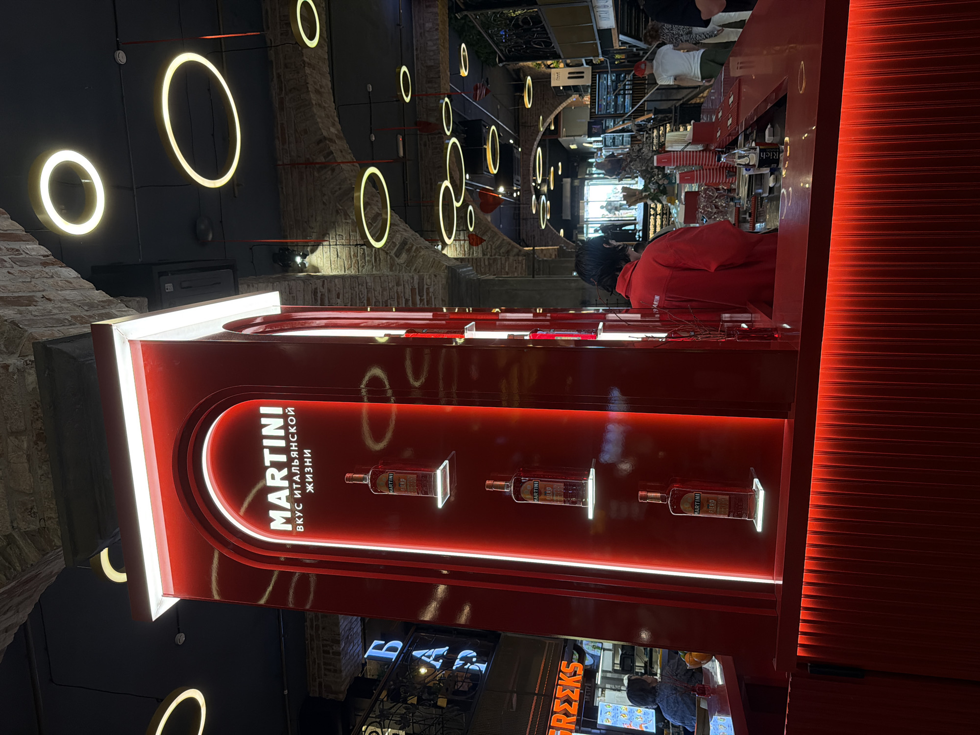 The image shows a bar area, likely in Moscow, Russia, given the Russian text on the Martini display, which translates to "Martini - Taste of Italian Life." The scene features a red, well-lit Martini display case showcasing several bottles of Martini Fiero on illuminated shelves. Above, decorative, circular lights are suspended from the ceiling. Behind the display, a person in a red shirt stands at the bar. In the background, other patrons and elements of the bar's interior are visible, including other bars. The environment suggests a modern, urban setting.