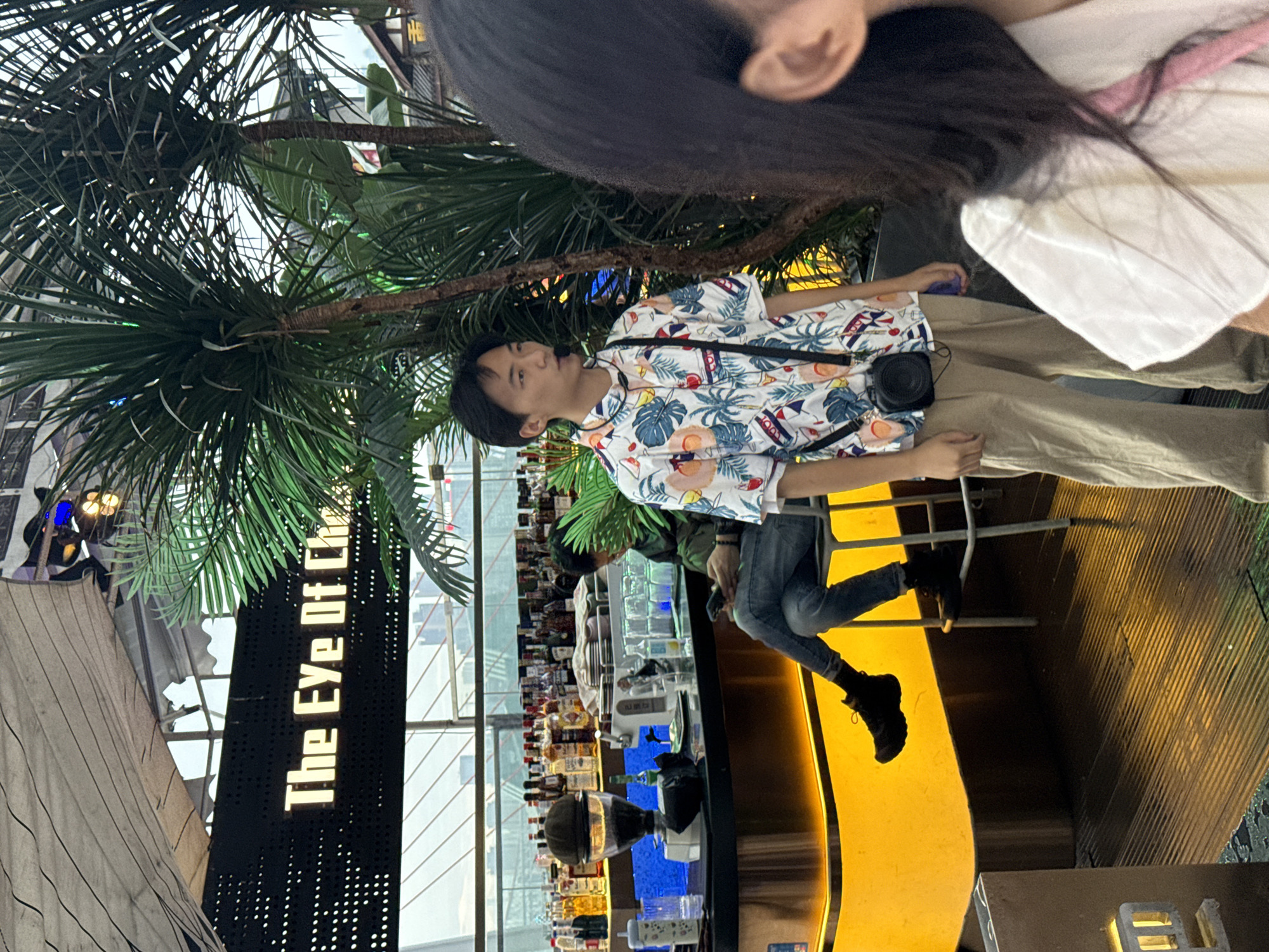An outdoor bar area, likely in Yuzhong District, China, is depicted with a bright yellow counter illuminated by LED lights. Behind the counter, numerous bottles of alcohol are displayed on shelves. A man in a black jacket and jeans sits on a stool, looking at his phone. In the foreground, a young man wearing a Hawaiian-print shirt and tan pants stands, holding a camera bag. To the right, the back of a person with long dark hair and wearing a white shirt is visible. Palm trees and lush foliage are integrated into the setting, suggesting a tropical or garden-themed atmosphere. The overall scene conveys a casual and vibrant ambiance.