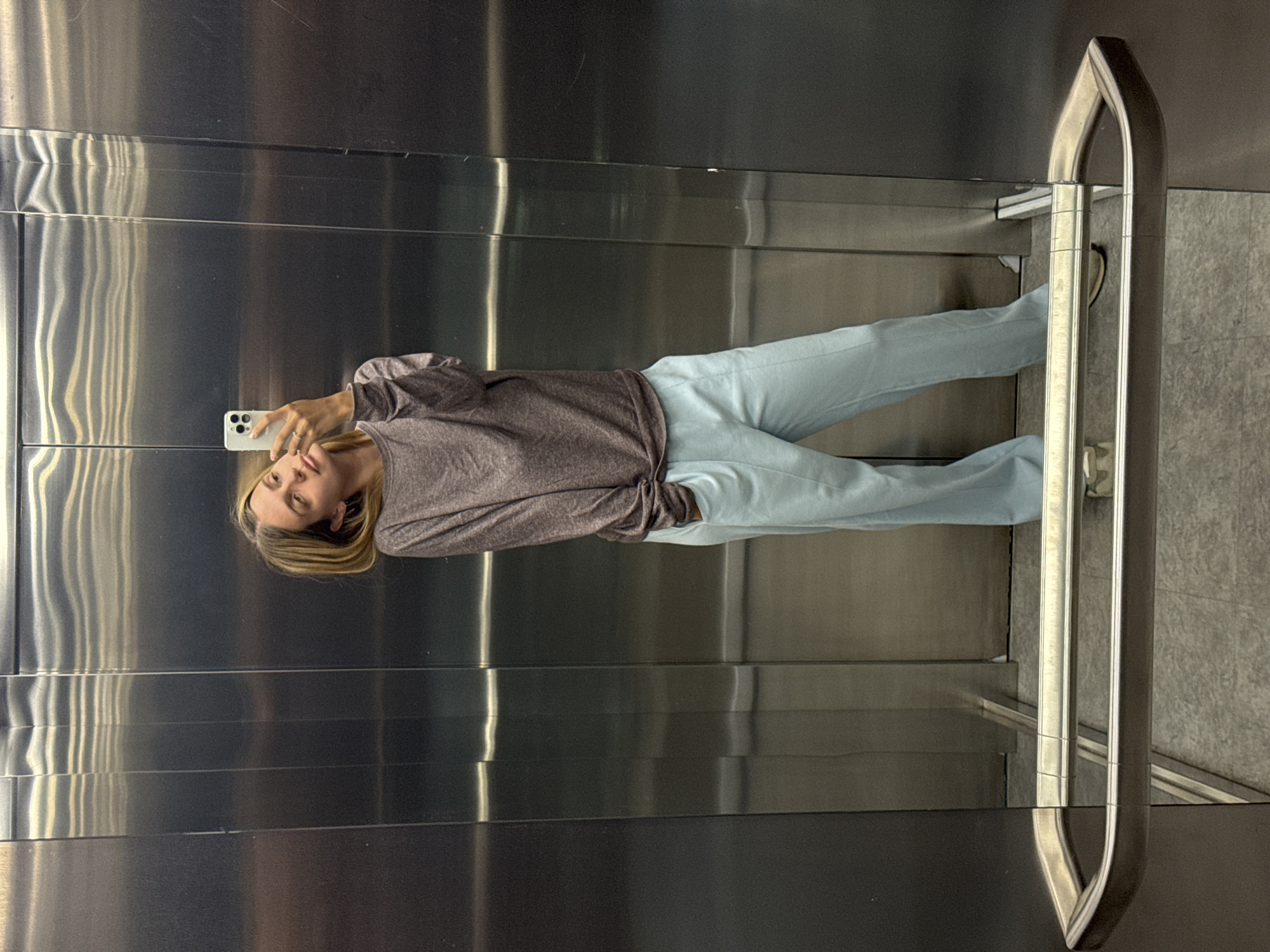 The image is a full-body mirror selfie taken by a young adult woman inside an elevator. The photograph is oriented vertically, showing the woman standing upright within the reflective space. She holds a white smartphone with a visible triple camera array in her right hand, pointed towards her, as she looks at the device. She has light-colored hair and wears a long-sleeved, brownish-grey top paired with light blue pants. A silver-colored ring is visible on her left hand.
The setting is an elevator characterized by highly polished, reflective metallic walls, which appear to be stainless steel. These walls show reflections of the woman and create vertical patterns of light and shadow, indicating the texture or panels of the walls. On the right side of the frame, a sleek, curved metal handrail is present. A portion of the elevator floor is visible in the bottom right, appearing as light-colored, possibly tiled, surfacing. The overall atmosphere suggests an indoor, modern environment. There is no visible text in the image, and the time of day is not discernible.