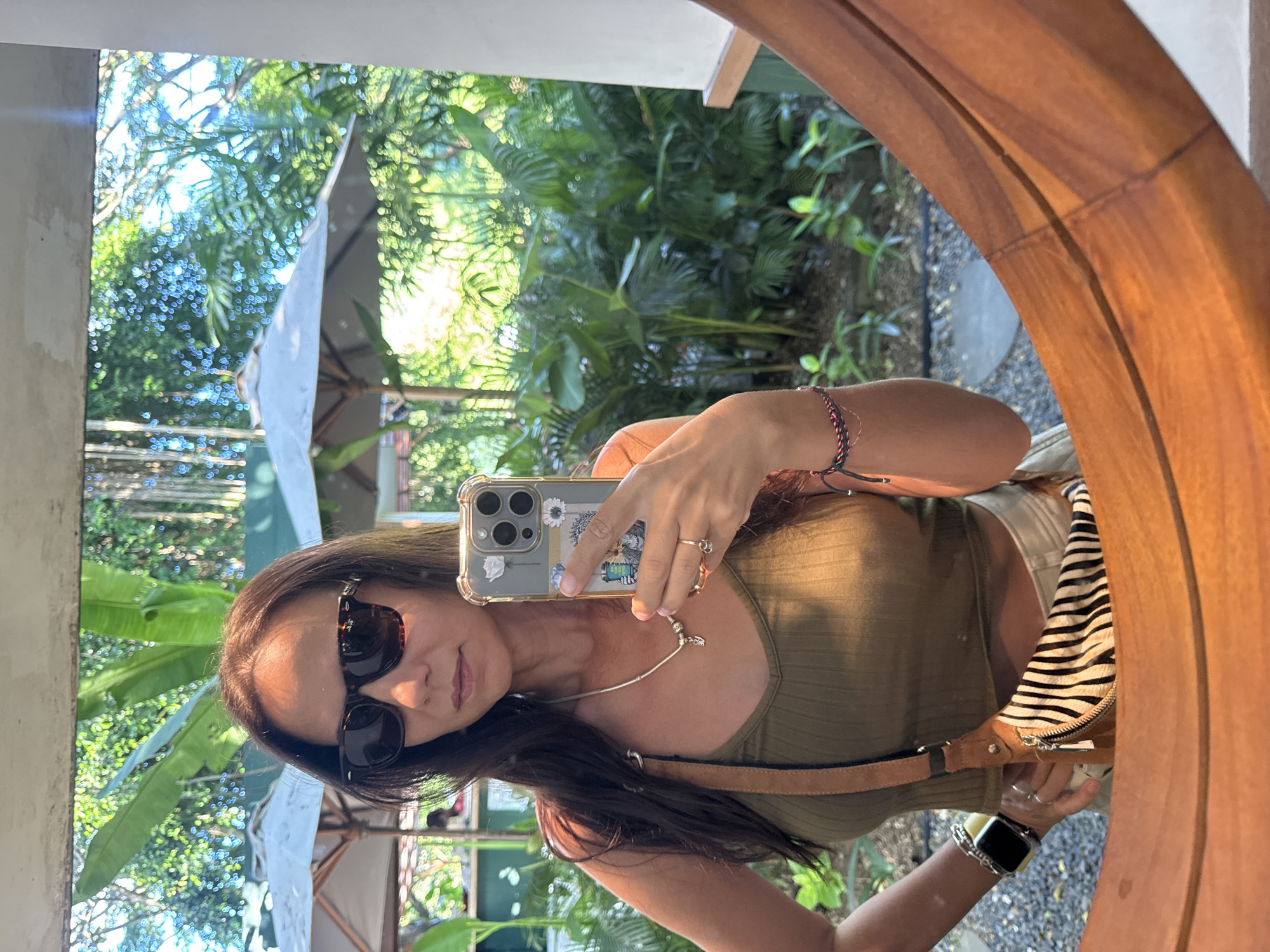 A woman is reflected in a round wooden-framed mirror, holding a smartphone to take a selfie. She wears sunglasses and an olive-green tank top with a brown leather shoulder strap. A zebra-striped handbag is slung over her shoulder. Her dark hair falls past her shoulders, and she wears jewelry, including a necklace, bracelets, and a ring.
The reflection behind the woman reveals a lush, tropical setting with greenery and parasols. The location is identified as Kuta Selatan, Indonesia, based on the vegetation and outdoor setting.
