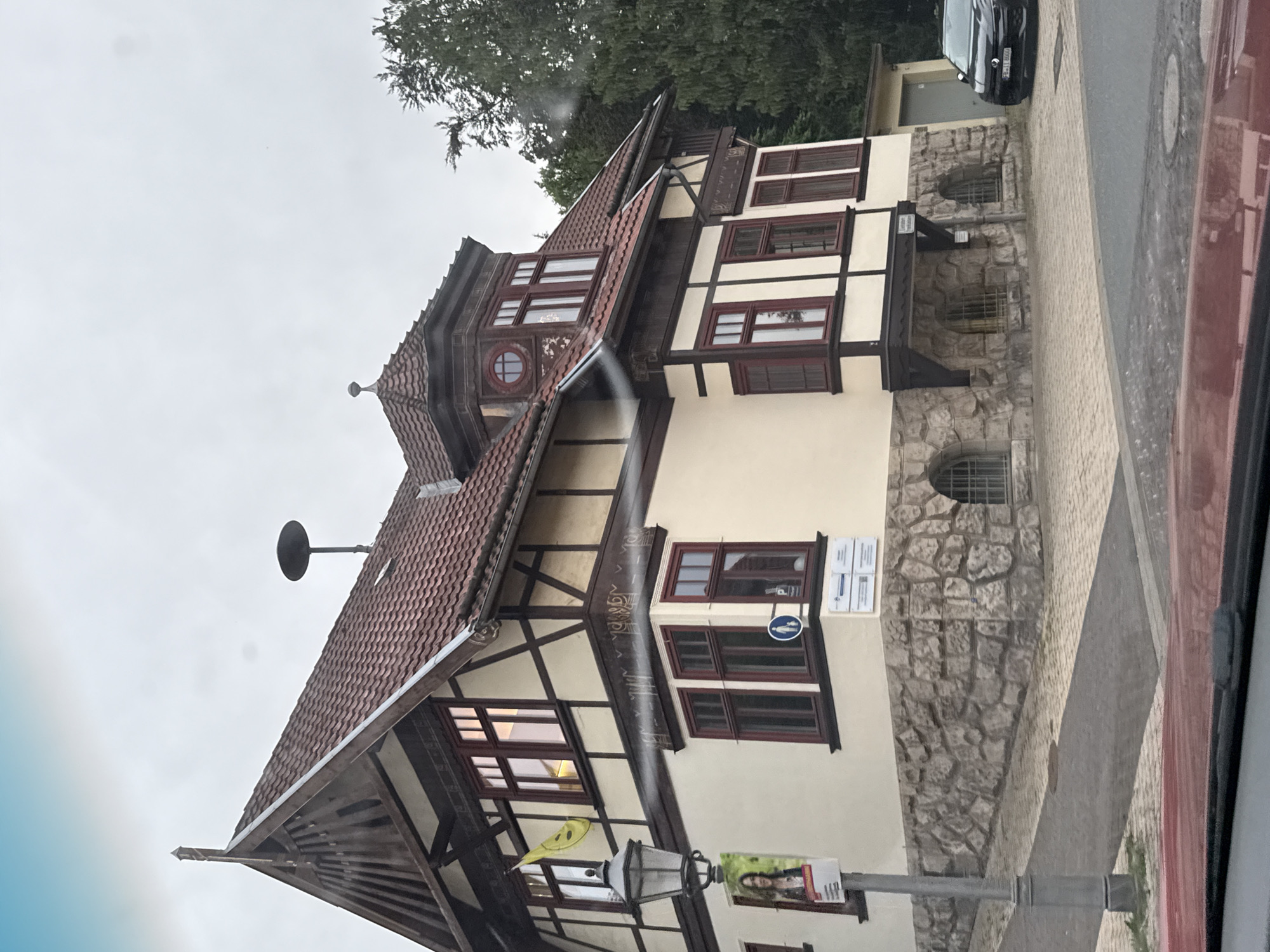 The image shows a two-story building with a red tiled roof and timber framing in the German town of Bad Sachsa, Germany. The building has cream-colored walls and dark red window frames. On the left side of the building, a lamppost stands next to a poster featuring a woman. A yellow flag with a smiley face is attached to the building. The lower portion of the building is constructed with rough-hewn stone. A dark-colored car is parked to the right of the building, which appears to be a residential or administrative building. The sky is overcast, and trees are visible in the background. The image seems to be taken from a vehicle.