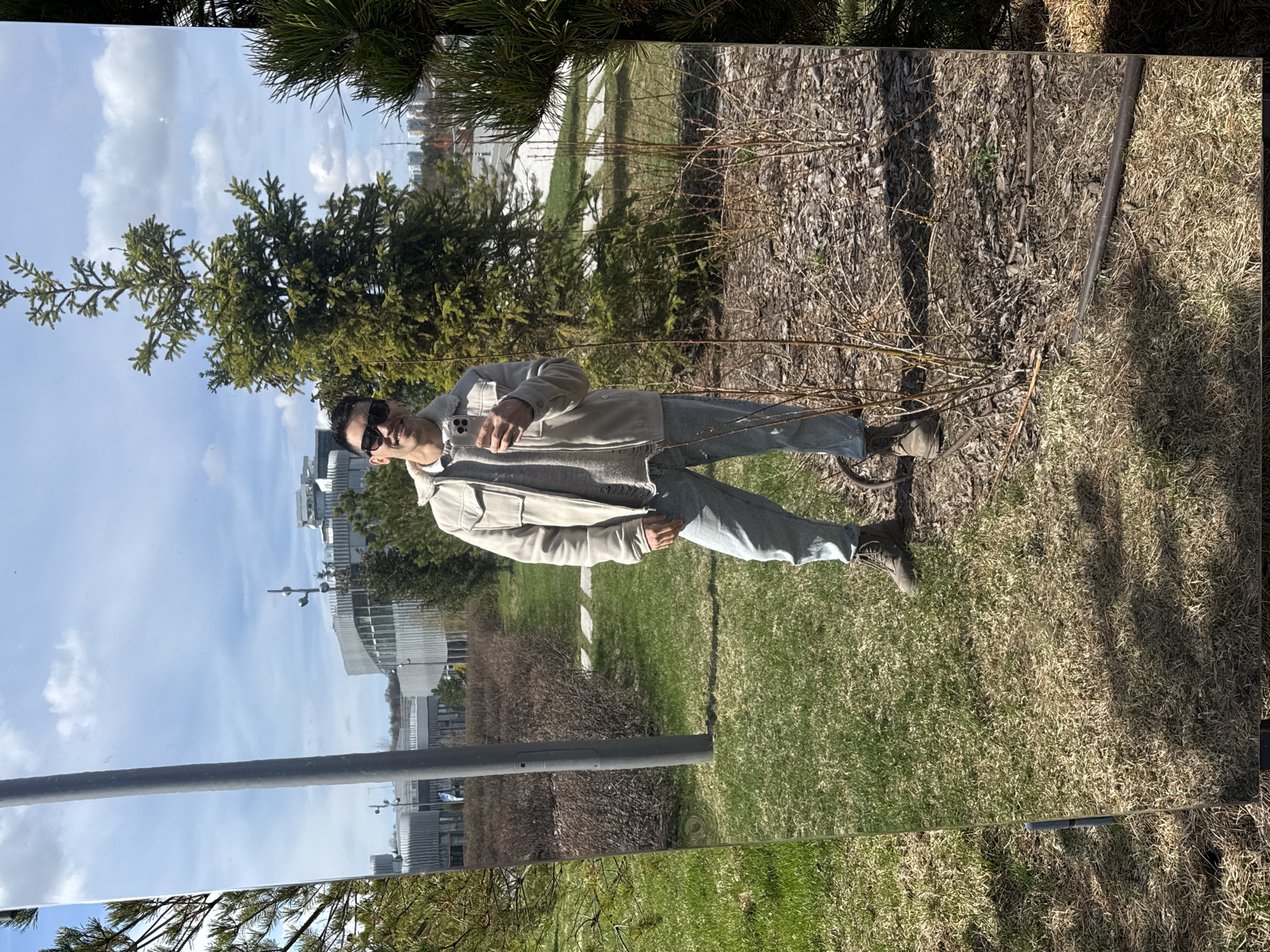 The image is an outdoor mirror selfie taken on a bright, sunny day with scattered white clouds. A man is reflected in a large, vertical, rectangular mirrored panel, which occupies the majority of the frame. He is standing upright, wearing sunglasses, a light-colored (possibly beige or off-white) jacket with a textured, shearling-like lining, light-wash jeans, and brown boots. He holds a smartphone in his right hand, apparently taking the photograph. His expression is mostly obscured by the sunglasses.
The reflective panel captures a detailed outdoor environment. In the background of the reflection, behind the man, are lush green pine trees against a clear blue sky. Distant, modern buildings with curved architectural elements are also visible in the reflection. The reflected ground shows green grass and paved paths.
In the actual foreground, outside the mirror's surface, the ground is covered with dry grass, wood chips, and bare branches of shrubs, suggesting a season like early spring or late autumn. A thick, dark pipe or structure lies on the ground near the right edge of the frame. A thin, dark pole is also visible vertically along the left side of the reflective surface in the reflection.
The scene depicts a public outdoor space, potentially a park or an area featuring art installations, located in Kazan, Russia. There is no visible text in the image.