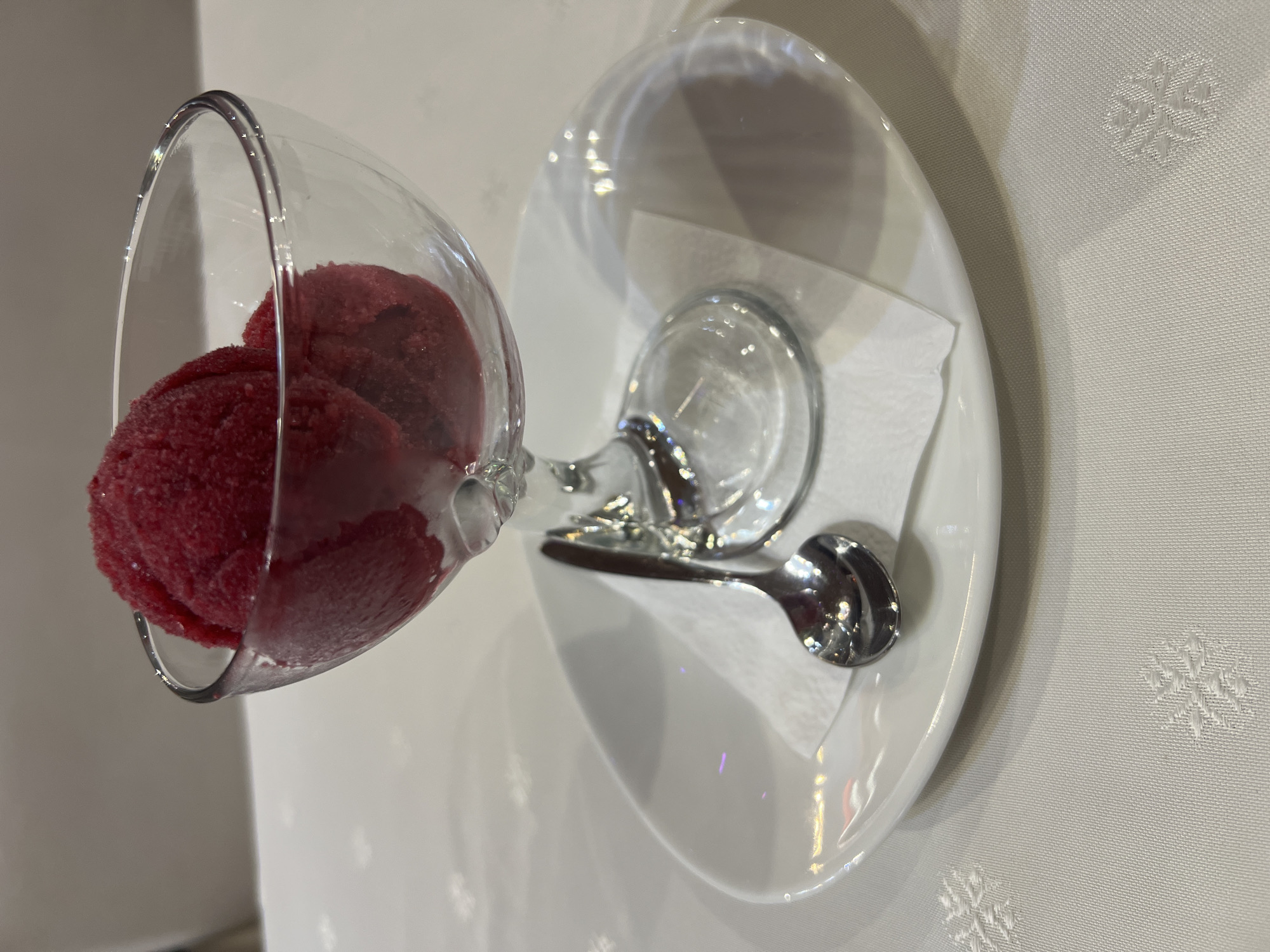 A close-up, eye-level shot shows a glass dessert bowl filled with two scoops of red sorbet, which is placed on a white cloth napkin on a white plate. A silver spoon rests on the napkin to the left of the bowl. The background is blurred, with a white tablecloth with a subtle snowflake pattern visible at the bottom. The scene is lit from above, creating soft shadows. The image is in focus and sharp. The location is Unknown, Unknown.