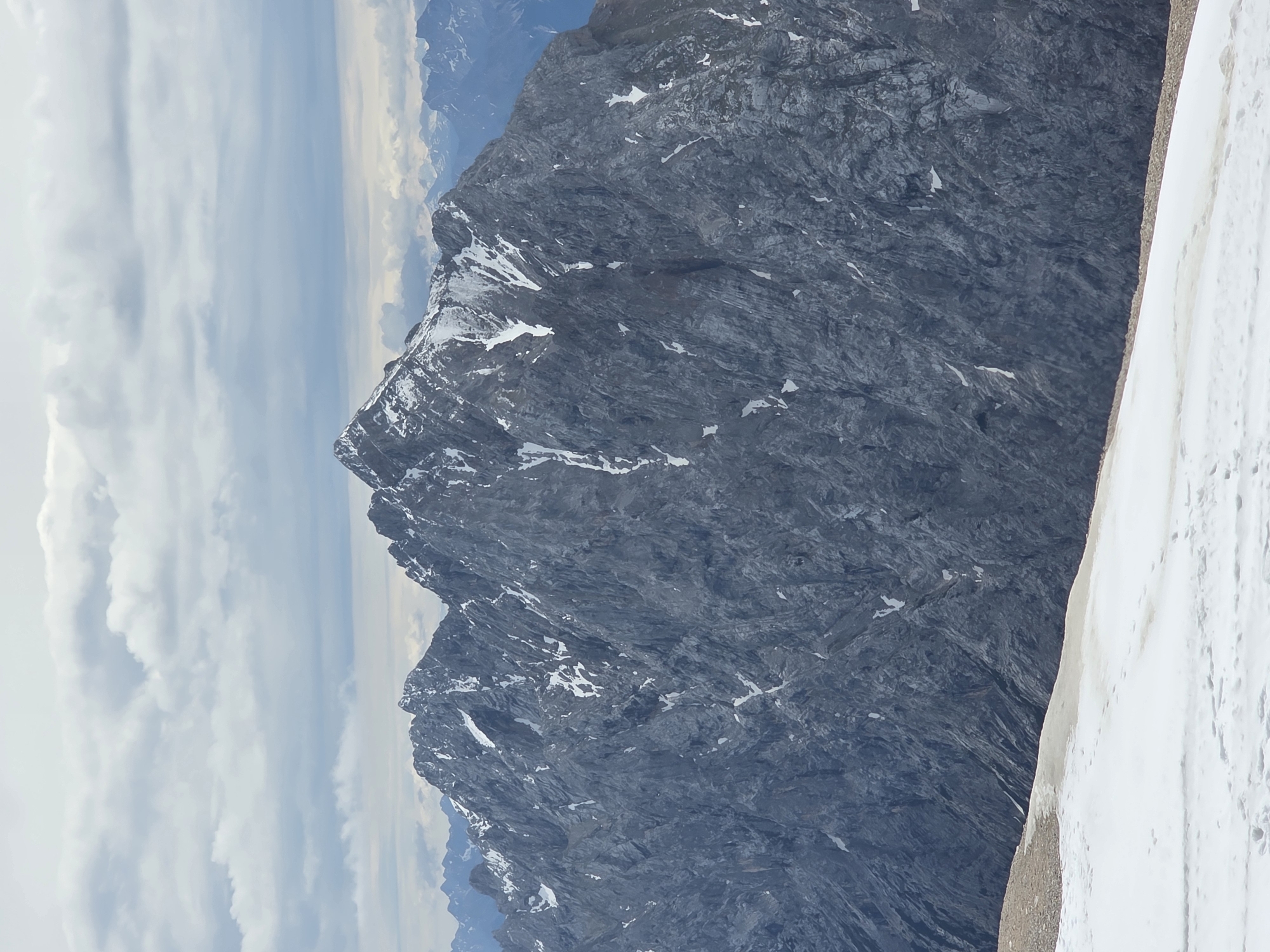 This image captures a wide, high-altitude mountain landscape on an overcast day, set in Unknown, Unknown. The central focus is a rugged, dark grey mountain range featuring multiple jagged peaks, heavily textured with exposed rock faces. Patches of white snow and ice cling to the crevices and slopes of these mountains. In the immediate foreground, a field of white snow with scattered brownish-orange discoloration covers the ground, transitioning to a strip of lighter brown, rocky terrain before meeting the base of the prominent peaks. The sky above is entirely covered by a thick layer of light grey and white clouds, suggesting diffuse lighting conditions. In the far distance, faint, hazy blue-grey mountain silhouettes indicate an extensive mountain range. No people, animals, or man-made structures are visible.