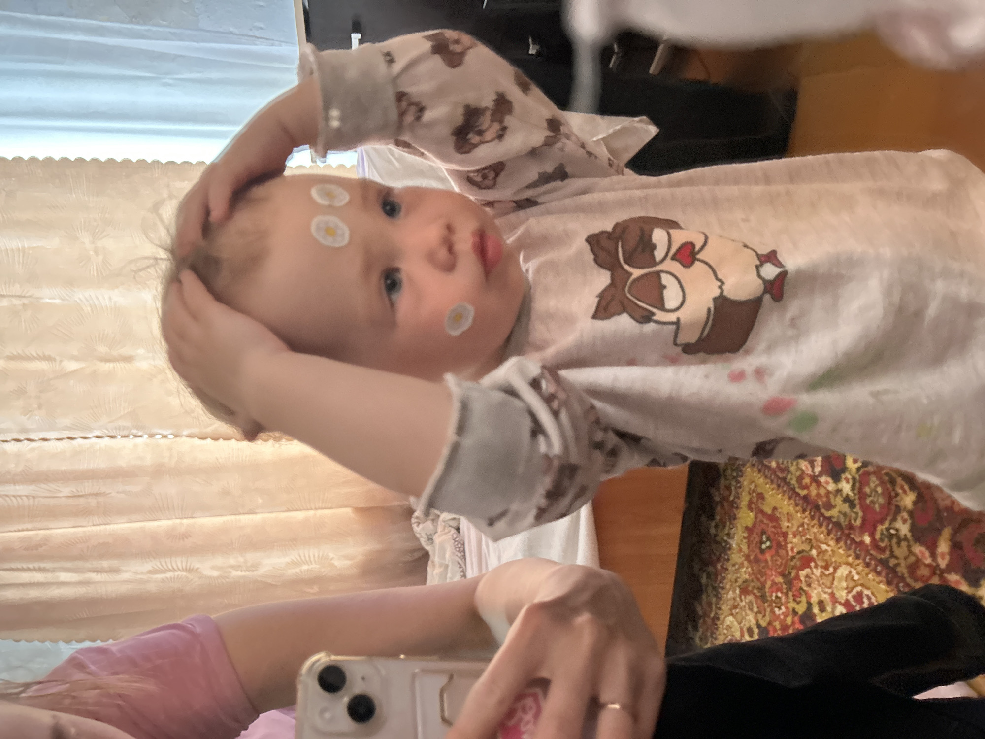 This image is a mirror selfie or reflection, primarily featuring a baby and parts of an adult. The baby, likely a toddler, has fair skin, light hair, and blue eyes, and looks directly at the camera with a neutral to slightly curious expression. Several white daisy-shaped stickers are visible on the baby's forehead and left cheek. The baby is dressed in a light-colored, long-sleeved onesie adorned with a charming brown cartoon owl character, complete with large eyes and heart details, printed on its chest. The baby's hands are raised and positioned near its head.
In the lower portion of the frame, an adult's arm and hand are visible. The adult is wearing a pink top and a simple gold ring on one finger. They are holding a light-colored smartphone, which appears to be an iPhone due to its distinctive triple camera lens array on the back.
The scene is set indoors, likely within a home in Азинский, Russia. The background includes light-colored curtains or blinds with a subtle pattern and a colorful, ornate rug featuring traditional red, gold, and green patterns. The indoor lighting suggests the photo was taken during the day or in a brightly lit evening setting. There is no visible text in the image.