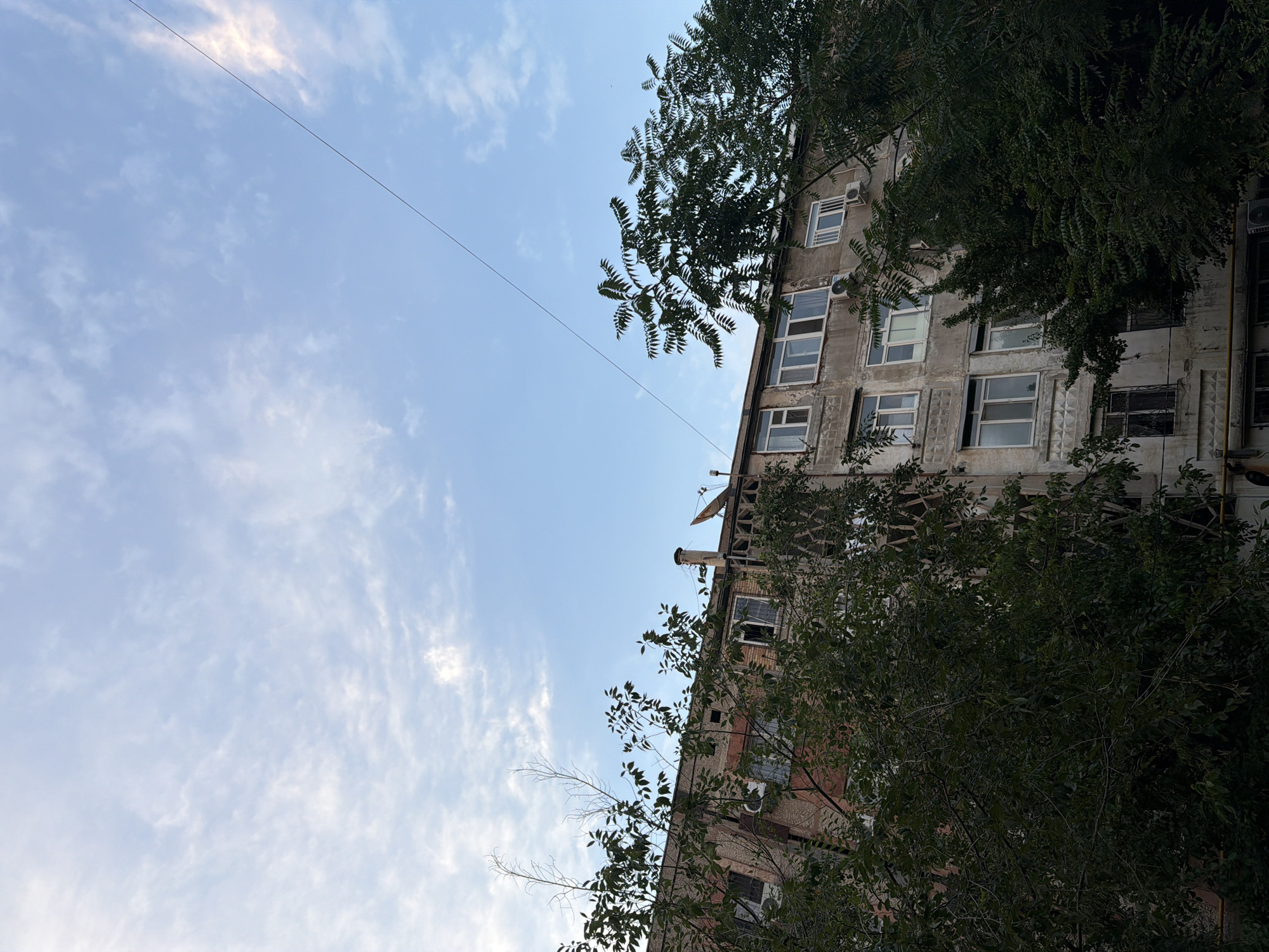 In Shymkent, Kazakhstan, a low-angle shot captures an aging, multi-story apartment building partially obscured by dense, dark green foliage. The building's facade is weathered, with a mix of visible brickwork and peeling plaster. Numerous windows, some with white frames, punctuate the structure, and several air conditioning units are attached to the exterior. Above the building, a vast, pale blue sky is dotted with wispy white clouds, and a thin, dark wire stretches diagonally across the frame. The scene appears to be an ordinary urban residential area, with no people or discernible activity visible.