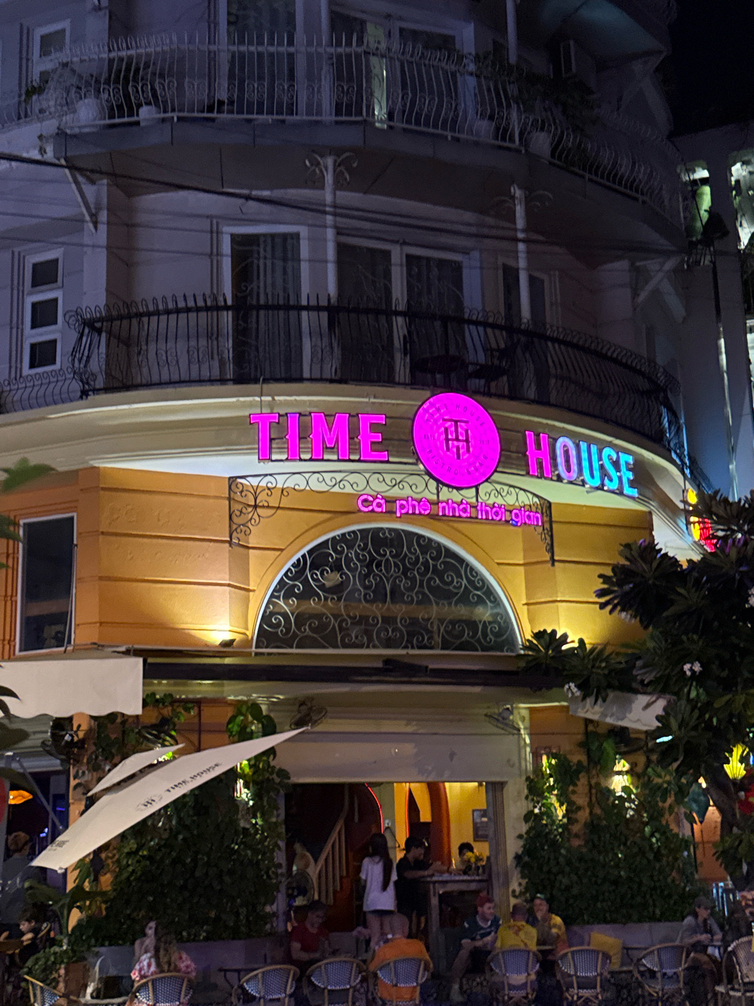 The image shows a street-level view of a building at night in Nha Trang, Vietnam. The building has multiple floors with balconies and ornate railings. A neon sign on the second floor reads "TIME HOUSE" in pink and blue lettering. Below the sign, a smaller pink neon sign says "Ca phe nha thoi gian," which translates to "Time house coffee." The ground floor appears to be a cafe or restaurant with outdoor seating and umbrellas. Several people are visible sitting at tables and walking around. The overall atmosphere is lively and suggests a commercial establishment operating at night. The architectural style and the presence of palm trees and tropical foliage are indicative of a coastal city in Southeast Asia like Nha Trang.