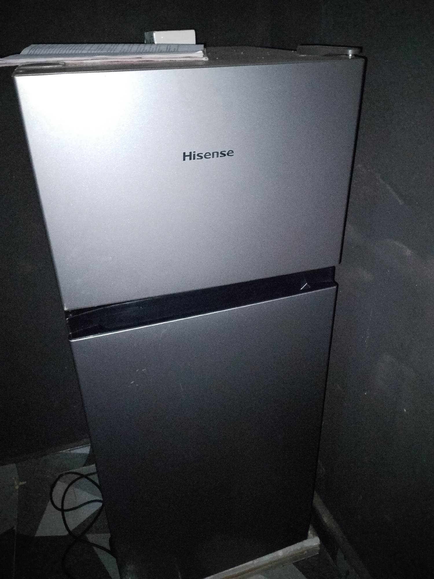 A Hisense brand refrigerator is shown in this medium shot, with its top half displaying the brand name prominently. The refrigerator is silver and stands in an indoor environment, possibly a kitchen or pantry, with a dark, textured wall behind it. On top of the refrigerator, a small stack of papers or manuals is visible, along with a white object that might be a power adapter. The lower portion of the refrigerator is darker, and a black power cord is coiled on the floor to the left of the appliance. The overall scene is dimly lit.