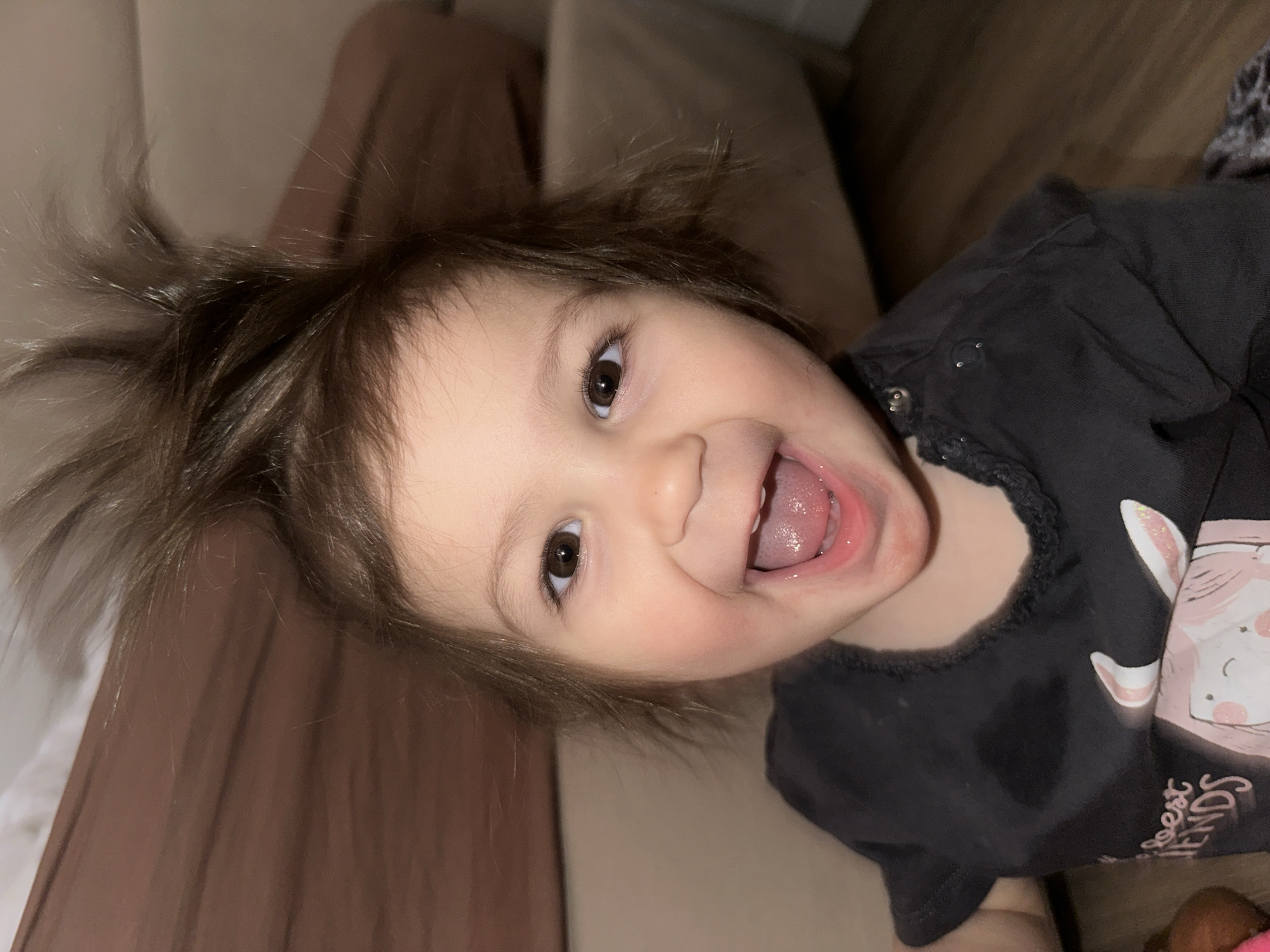 A young child with brown hair is smiling widely with their mouth open, revealing their tongue and teeth. The child is wearing a dark shirt with a cartoon graphic. The background consists of a couch with a beige or brown hue. The image does not contain sufficient information to verify the location in the city of Khimki, Russia.