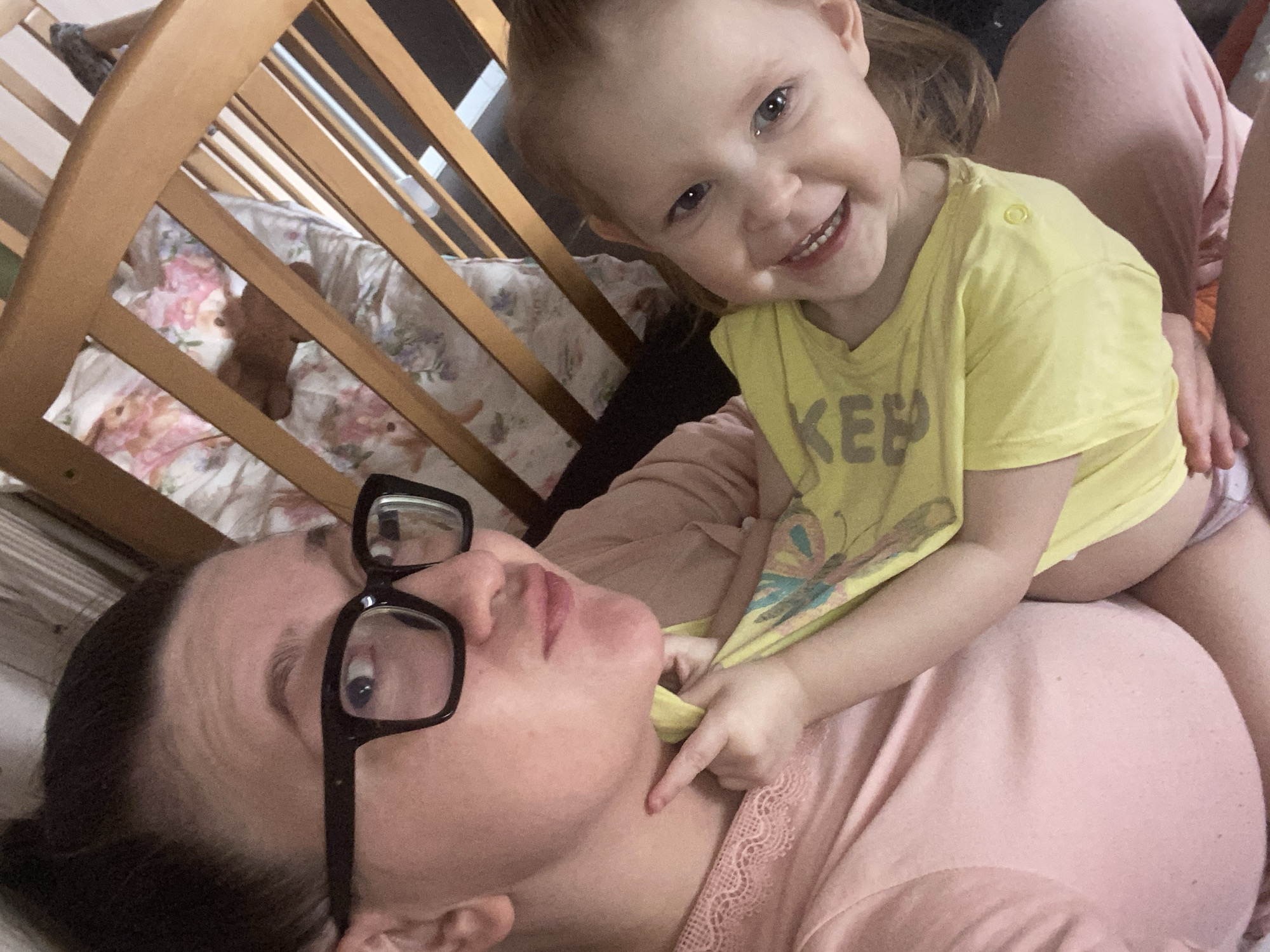 The scene is an indoor environment in Chaikovsky, Russia. Two individuals, an adult female and a young child, are depicted in a close-up, likely self-taken photograph. The adult female, with dark hair tied back, wears black-rimmed glasses and a light pink top with lace detailing. She poses with pursed lips, looking towards the camera. A young child, appearing to be a toddler with light red hair, wears a yellow t-shirt with partial text "KEE" visible and light-colored bottoms. The child is smiling broadly, looking directly at the camera. The child's right index finger points towards the adult's neck, while the child's left hand grasps and pulls up her own yellow t-shirt. Both individuals are in close physical contact, with the child positioned in front of or on the lap of the adult. In the upper right background, a light brown wooden crib with vertical slats is visible. Inside the crib, bedding with a floral pattern (pink, green, white) and a brown plush toy are discernible.