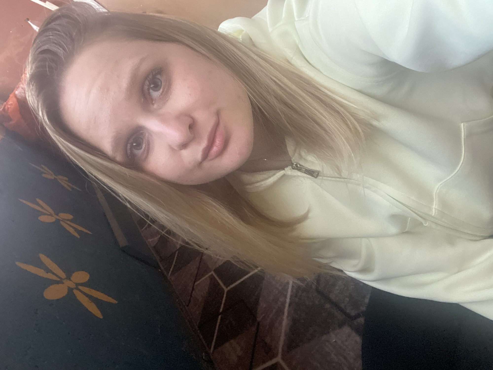 The image appears to be a selfie of a blonde woman wearing a light-colored hooded sweatshirt. She is reclining on a surface with a dark fabric featuring yellow floral or foliate designs. The background contains a patterned surface which looks like a wooden floor. It is not possible to verify if the location is Chaikovsky, Russia from the image alone. There is no additional information visible regarding location.