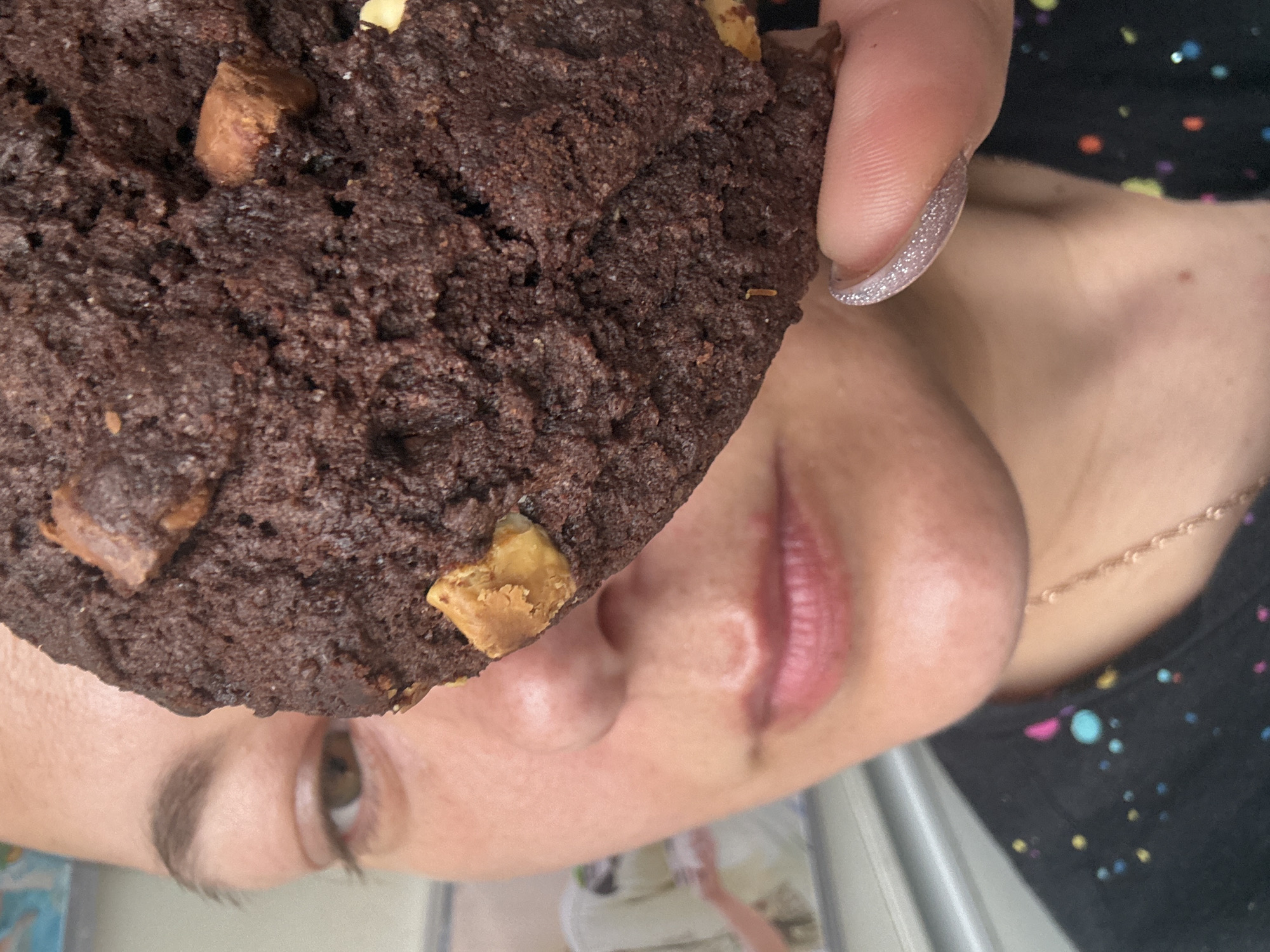 A vertically oriented, close-up photograph captures a person holding a large, deeply colored chocolate cookie directly in front of their face. The cookie, which dominates the foreground, has a rich, textured surface and is generously embedded with various chips and chunks, including dark chocolate and lighter, possibly peanut butter or white chocolate pieces. The person's fingers, holding the cookie, are visible, with one fingernail featuring a sparkly, light pink or lavender glitter polish.
The lower portion of the person's face is visible in the background, angled from the bottom right of the frame. This includes an open eye with a visible pupil, the bridge of the nose, and slightly parted lips, suggesting an imminent bite or the enjoyment of the cookie's aroma. A delicate gold chain necklace is draped around their neck. The person is wearing a dark top adorned with a vibrant, multi-colored splatter pattern in shades of pink, blue, yellow, white, and orange.
The activity appears to be the act of preparing to eat or savoring a cookie. The setting is indoors and seems casual, with an indistinct light-colored background. No specific time of day or weather details are discernible from the visual information. The image conveys a sense of immediate pleasure or anticipation. There is no visible text within the photograph. The location context for this image is Dmitrov, Russia.