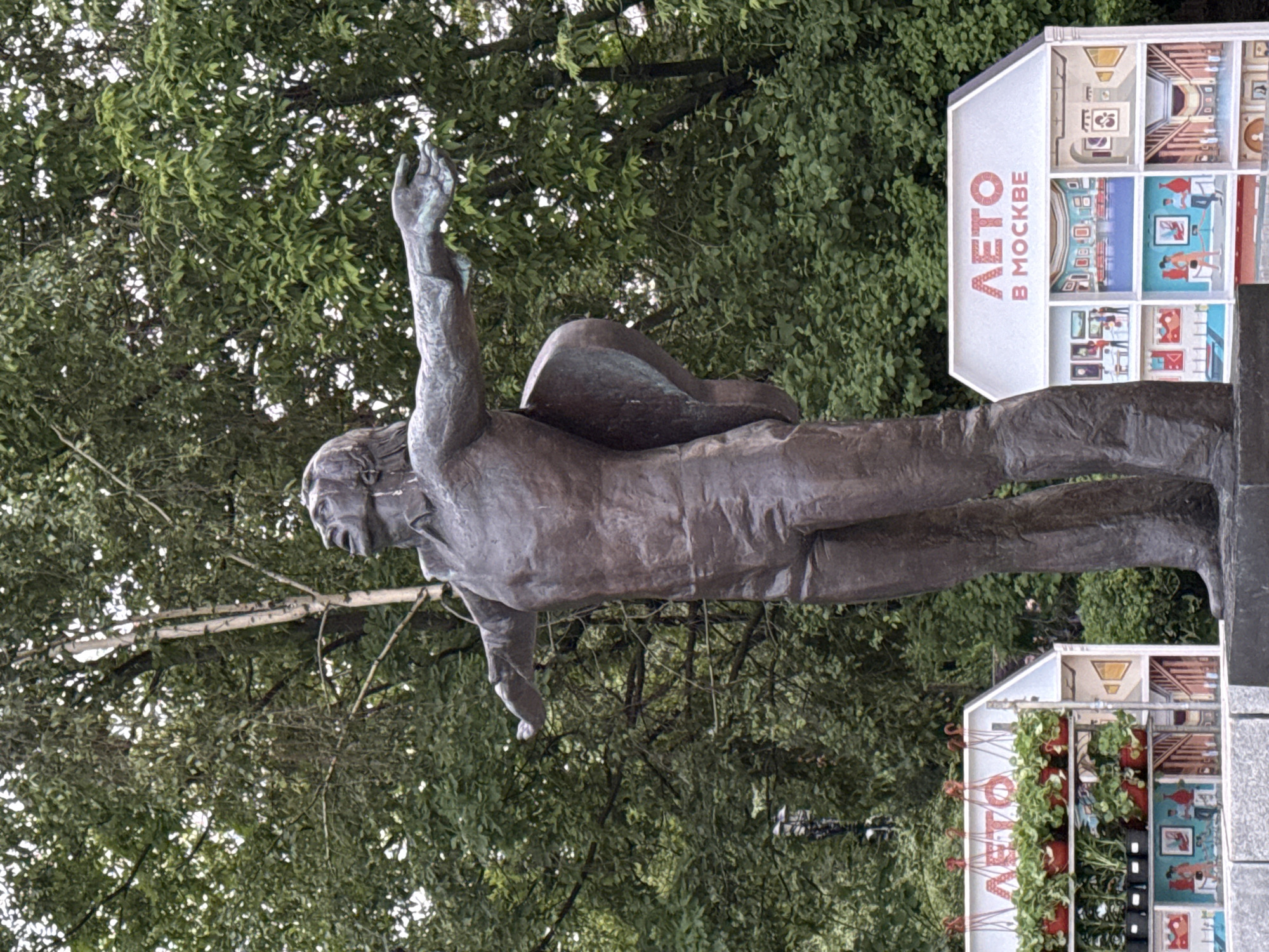 The image depicts a bronze statue of a man with his arm raised, seemingly in a gesture of greeting or acknowledgement, set against a backdrop of dense green trees. The man is wearing a suit and has what appears to be a satchel or bag slung across his back. Adjacent to the statue is a white sign with the words "ЛЕТО в МОСКВЕ" (Summer in Moscow) displayed prominently in red letters. The sign also features illustrations of various art gallery exhibits arranged in a grid. Below the sign, hanging plants are visible in small pots, indicating a decorative or landscaped area. The statue and sign are in an outdoor public space, likely a park or square in Moscow, Russia. The scene suggests a celebration or promotion of cultural events, possibly art exhibitions, during the summer months.