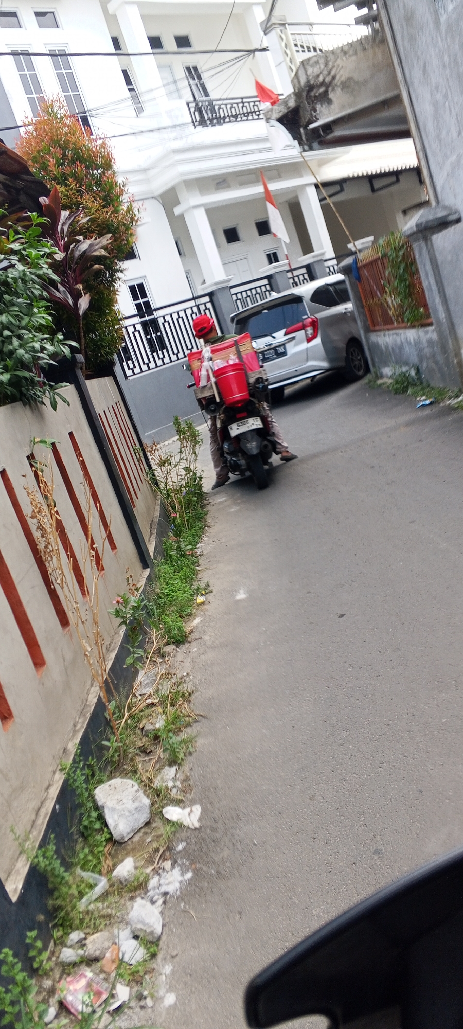 The image depicts a street scene in Depok, Indonesia, with a focus on a person on a motorcycle. A rider wearing a red cap and a green shirt is seated on a black motorcycle. The motorcycle is equipped with a large red box on the rear, suggesting it's used for deliveries. The rider is partially obscured by the motorcycle and its cargo.
To the left of the motorcycle, a modern white building with multiple windows and balconies is visible. Indonesian flags, red and white, are displayed on the building, indicating a national celebration or observance. A lush green tree with red leaves is positioned in front of the building.
The road is paved and appears to be a narrow street or alleyway. To the immediate left of the road is a concrete wall with decorative vertical orange stripes. A strip of vegetation, including small plants and dry branches, runs along the base of the wall. On the right side of the road, another building with a brown wooden fence is partially visible. The overall impression is a residential or commercial area in Depok.