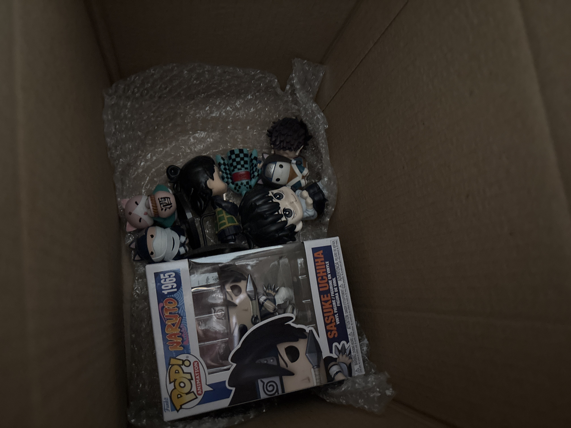 A cardboard box is filled with anime merchandise. Bubble wrap lines the interior of the box. Among the contents are several chibi-style anime figures and two Funko Pop boxes featuring "Naruto" and "Sasuke Uchiha."