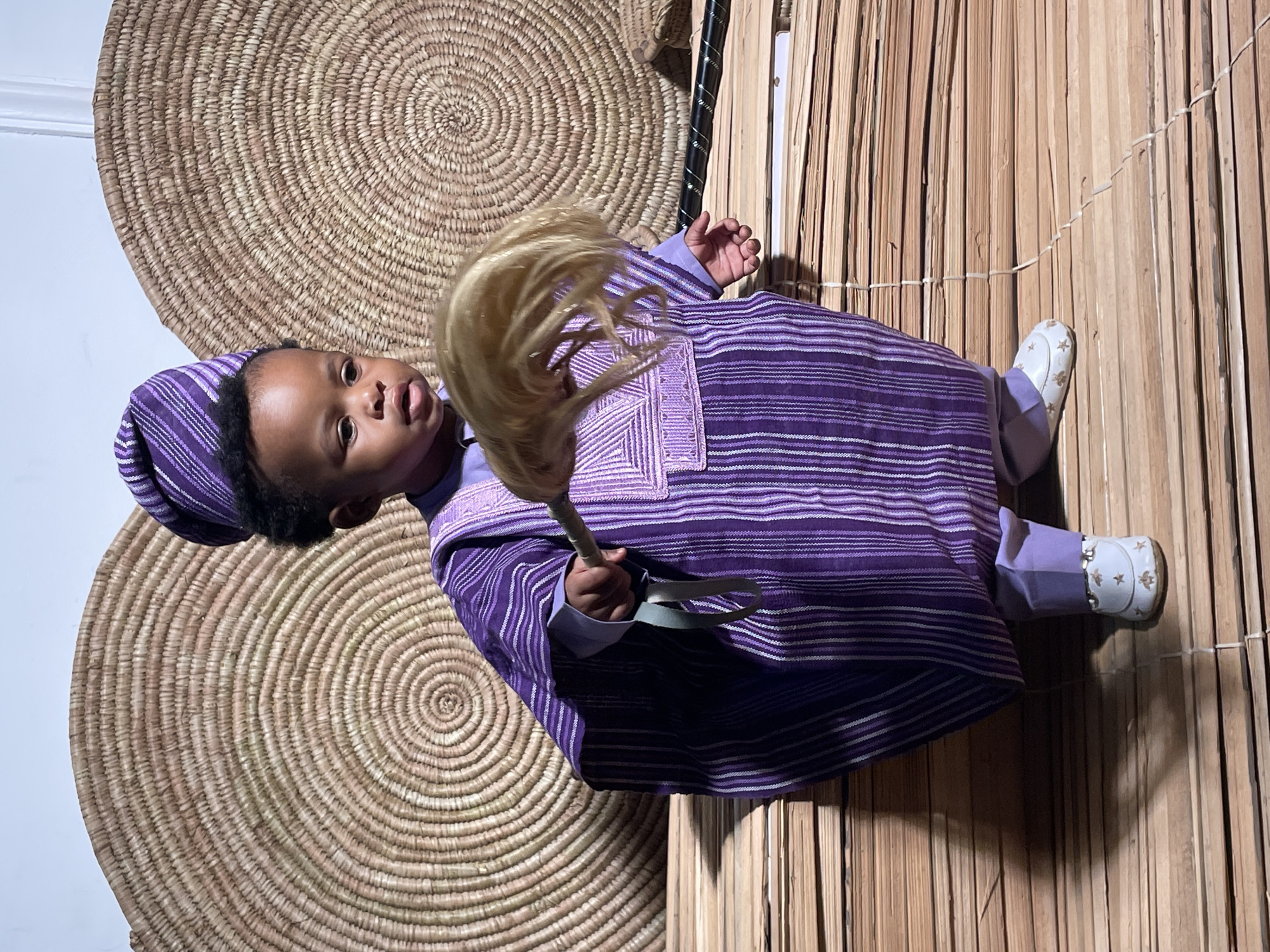 A baby in a purple Yoruba traditional outfit stands in front of a straw mat wall and a large flat woven basket, holding a horsehair fly whisk. The baby wears a matching purple cap and white shoes. The environment appears to be a studio setup, possibly in Jos, Nigeria, where Yoruba culture is present.
