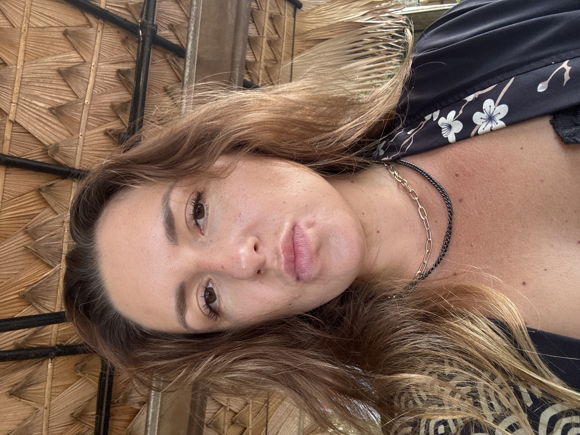 A woman is captured in a close-up, selfie-style portrait from the chest up, seated outdoors under a thatched roof. The woman has fair skin, brown eyes, and long, wavy brown hair. She is wearing a black garment with a floral pattern and two necklaces. Her lips are pursed, and she has a small blemish on her chin. The background consists of the textured, woven pattern of the thatched roof and a black support beam. The lighting suggests it is daytime.