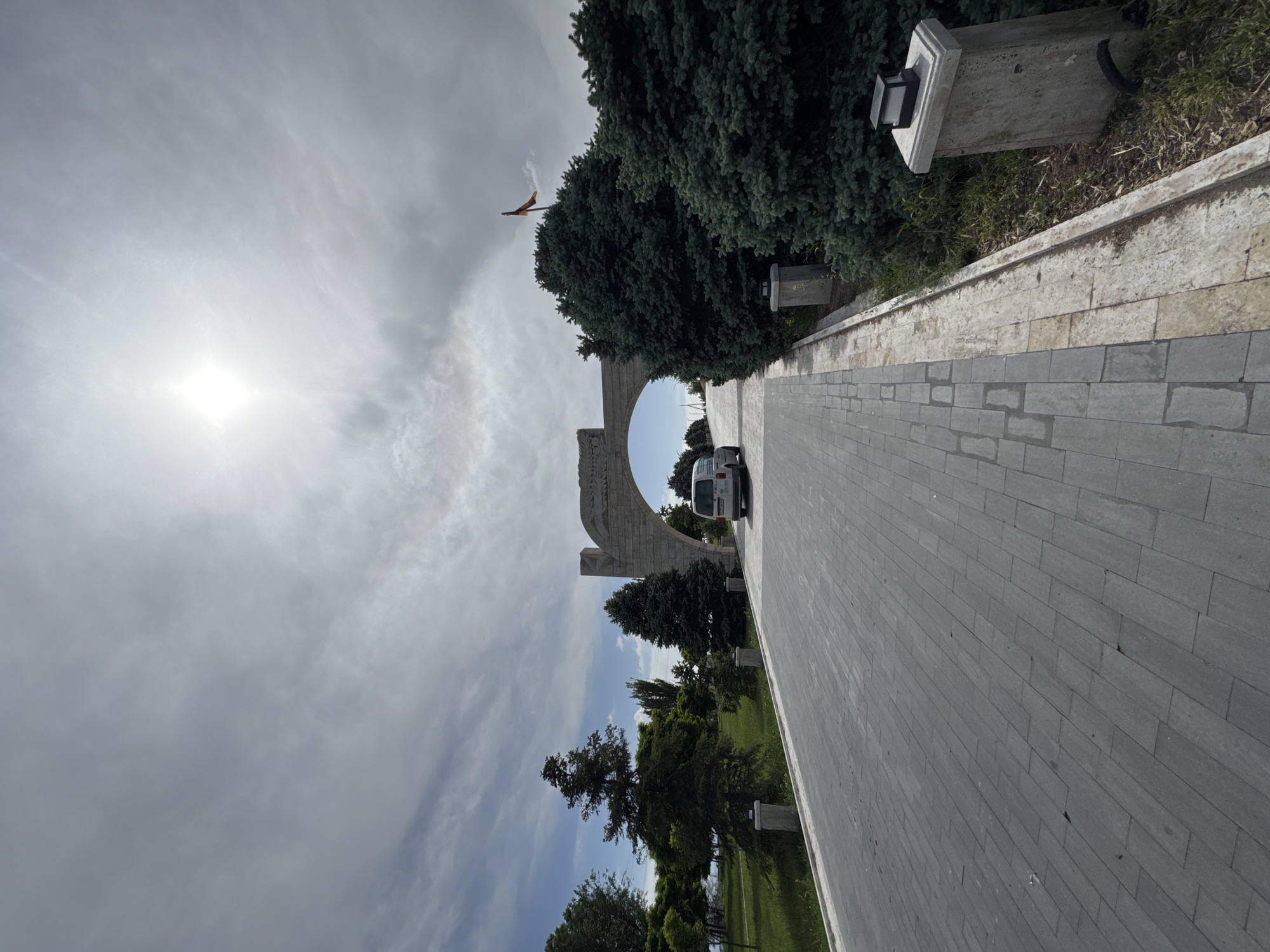 A street view in Yerevan, Armenia, features a grey brick path leading to an arched monument. A white van is parked beyond the arch. To the right of the path, there's landscaping with trees and decorative stone posts. The sky above is cloudy with the sun visible. A flag flutters atop the monument.