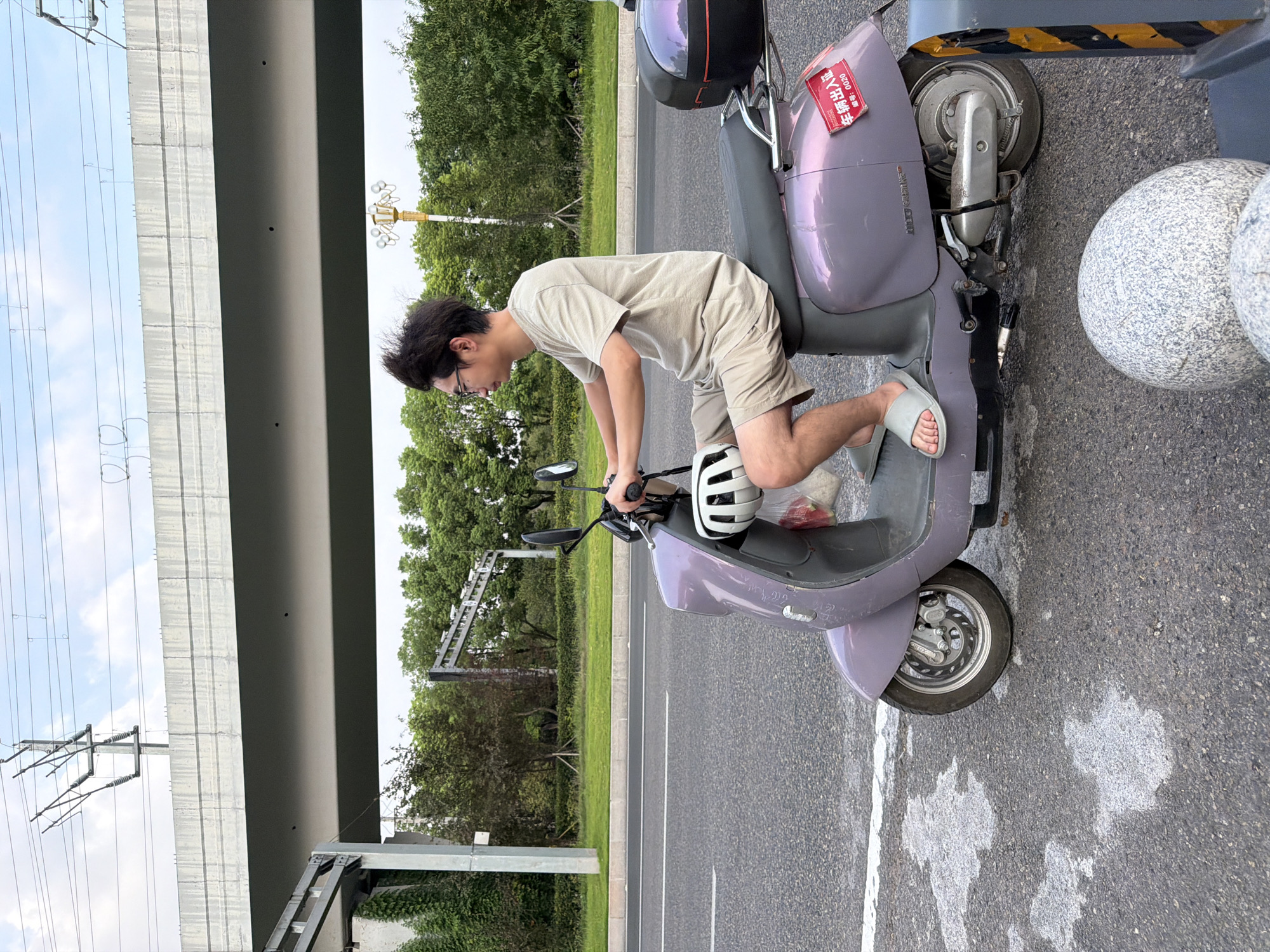 In Jiangjin, China, a young man with dark, tousled hair and glasses is riding a purple electric scooter on a paved road. He is wearing a light-colored t-shirt and beige shorts, with light gray slides on his feet. A white helmet is secured to the front of the scooter, and a red sticker with Chinese characters and the number "0020" is visible on the rear fender. The scooter has a black storage box mounted at the back. The road has white lane markings and some white patches that appear to be spilled material. In the background, a concrete overpass with overhead power lines stretches across the frame. Lush green trees and a bright sky with some clouds complete the scene.