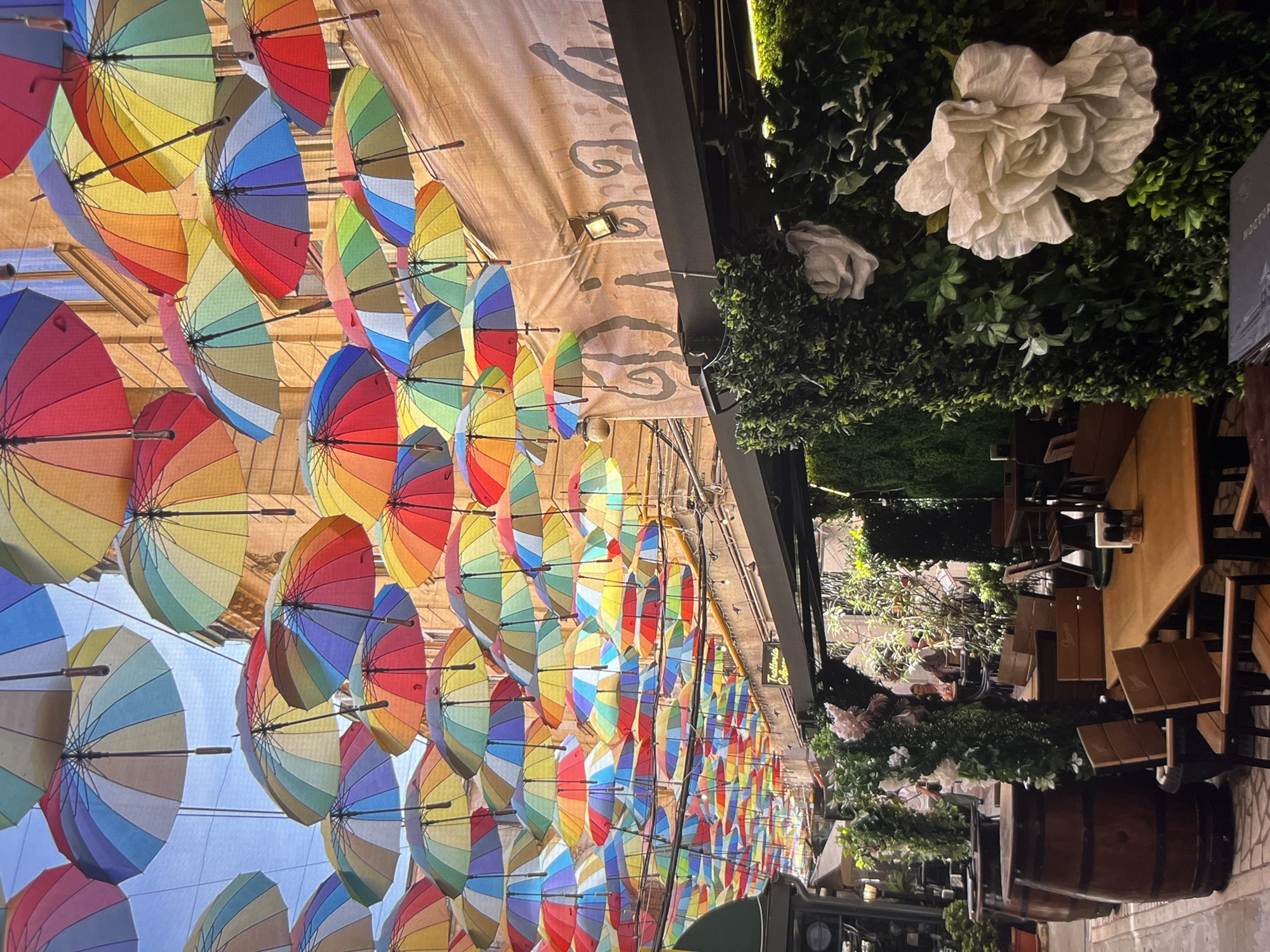 The image shows an outdoor cafe in an urban environment. Numerous brightly colored, rainbow-striped umbrellas are suspended overhead, creating a canopy above the cafe. The umbrellas are positioned to provide shade and add an artistic element. The cafe's structure features a tan awning with text in an indiscernible script. Plants with artificial white flowers adorn the space beside the awning. The cafe includes wooden tables and chairs arranged in a dining area, with people seated at some tables. A large wooden barrel is placed near the front of the cafe. The setting appears to be a narrow street or pedestrian area, suggesting a bustling urban location. The image was taken in the city of Unknown, Unknown.