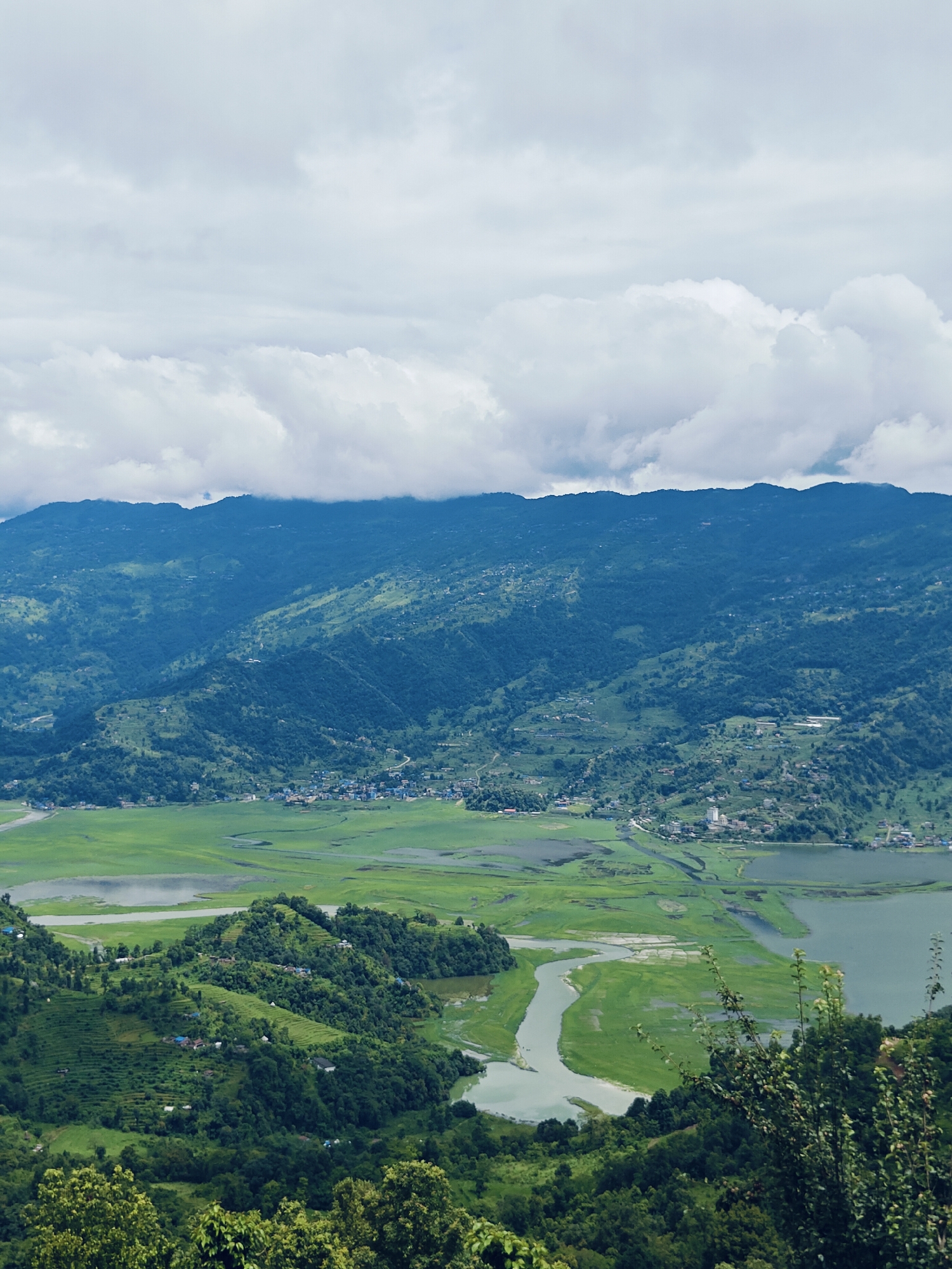 This is an aerial, high-angle, wide shot of a lush green valley in Pokhara, Nepal. The scene is dominated by rolling hills covered in dense vegetation and rice paddies. A winding river or canal snakes through the valley, leading to a large body of water on the right. In the middle ground, a small village with clustered buildings is visible on the side of a hill. The background features a mountain range, partially obscured by clouds. The sky is overcast with dramatic cloud formations. No people are visible in the image.