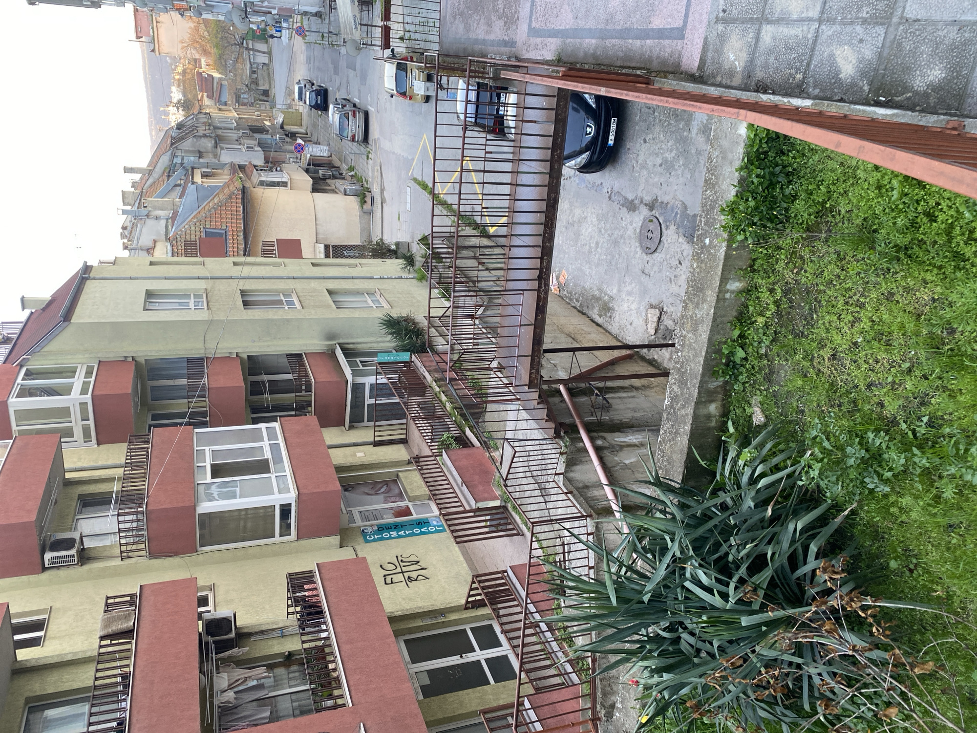 The image shows an exterior view of a multi-story building in Varna, Bulgaria. The building is painted in a combination of beige and red-brown, with each apartment having a balcony. External metal staircases provide access to the apartments. A sign on one of the floors says "DENTIST-STOMATOLOG", which indicates that one of the commercial tenants of the building is a dental office. A black car is parked on the street. The scene is shot from an elevated position looking down. The street is visible in the background with other buildings. A concrete barrier and some greenery are in the foreground. The weather appears overcast.