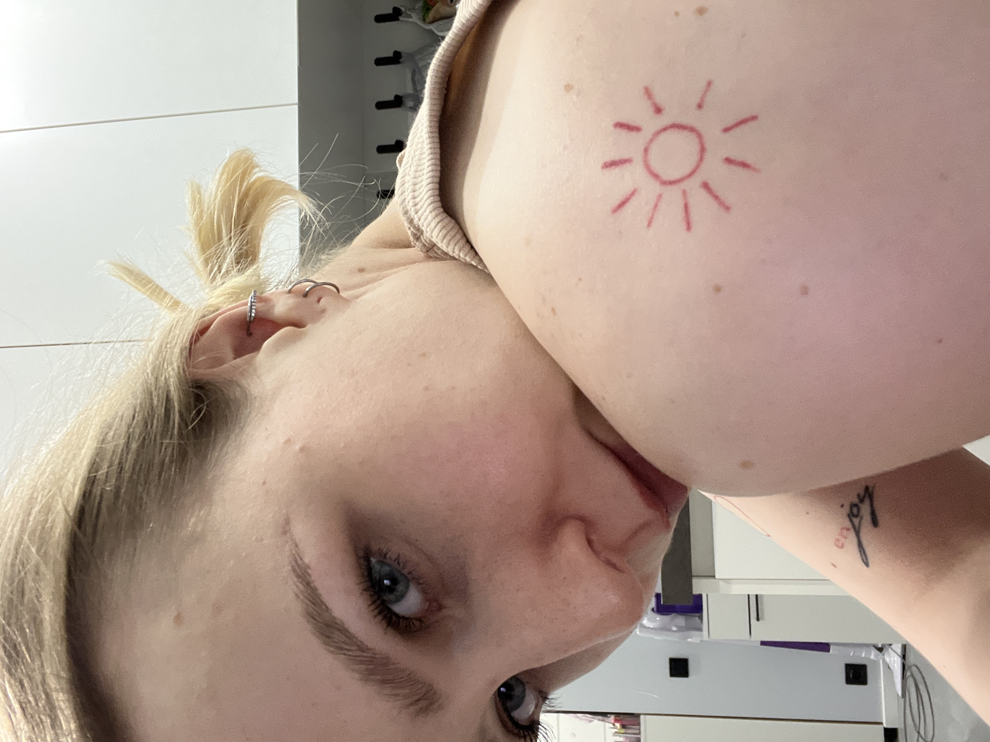 A close-up, rotated view of a fair-skinned young woman with light blonde hair partially pulled back in a ponytail. She has blue eyes and minimal makeup. There is a small silver earring visible in her left ear. A red, sun-shaped tattoo is present on her left shoulder. The tattoo is a simple line drawing of a circle with radiating lines. A portion of a beige-colored shirt or tank top is also visible. An "enjoy" tattoo is on the subject's left arm. The background includes a white wall with horizontal lines, and glimpses of a desk with papers and miscellaneous items. The image does not contain any location information, thus it does not include the location of Khimki, Russia.