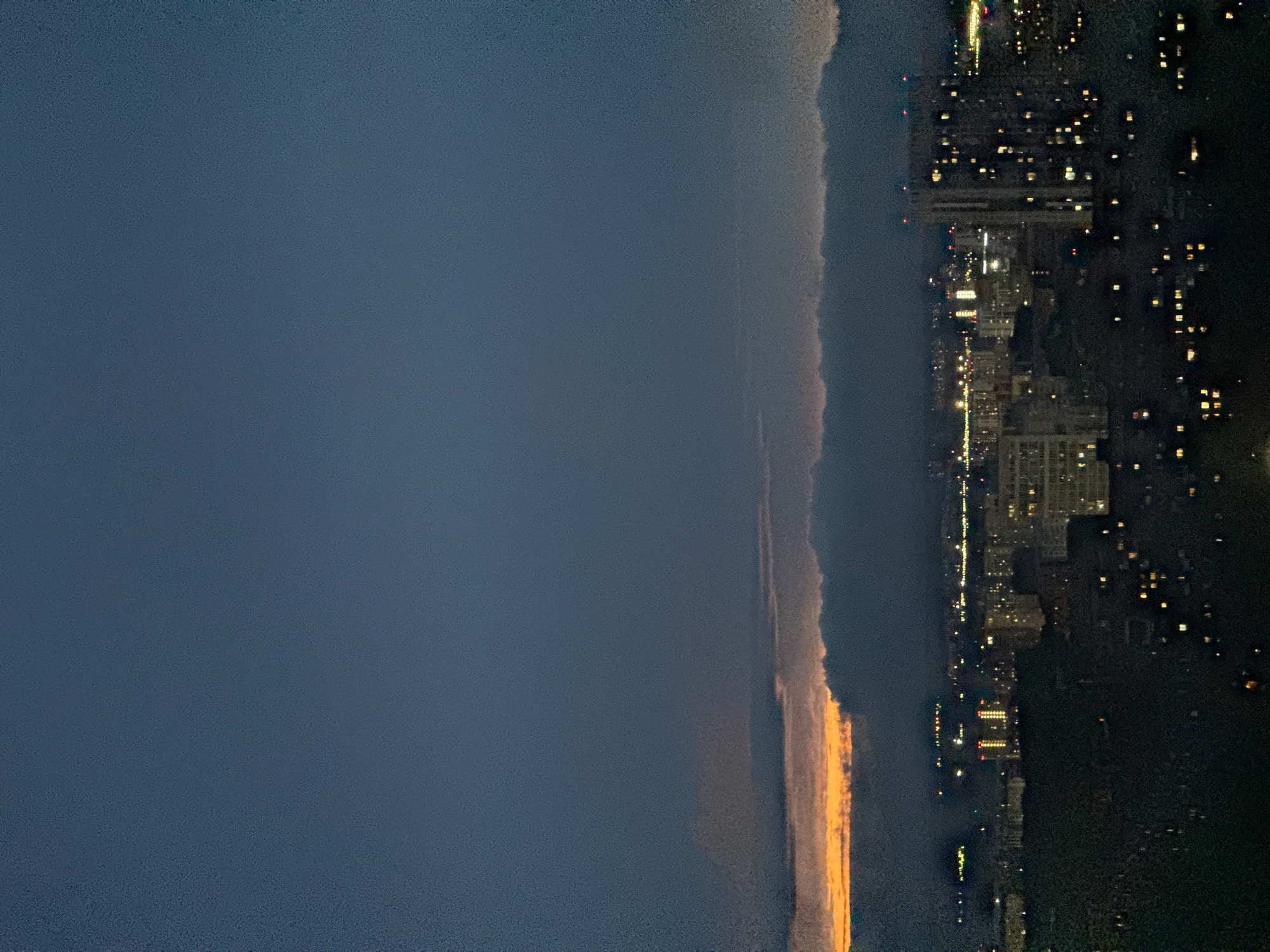 The image shows a nighttime view of what appears to be a coastline with city lights, possibly Гамово, Russia, bordering a body of water or open space. The sky is dark, transitioning into a lighter hue closer to the horizon where the cityscape is situated. Lights from buildings and infrastructure are visible, creating a scattered pattern across the dark land. A faint, vertical orange and gray band bisects the image. The left side of the image is largely dark, suggesting a vast expanse of sky or sea. The image is oriented sideways.