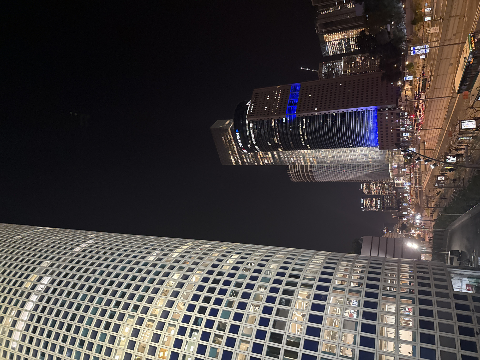 At night, the image captures a section of Tel Aviv, Israel. The foreground features a portion of the curved, grid-like façade of the Azrieli Sarona tower, characterized by its pattern of squares with visible interiors. Above and slightly behind this structure, several skyscrapers rise, their lit windows illuminating the night. The street scene is visible, showing a road and some pedestrians. There is some light in the sky.