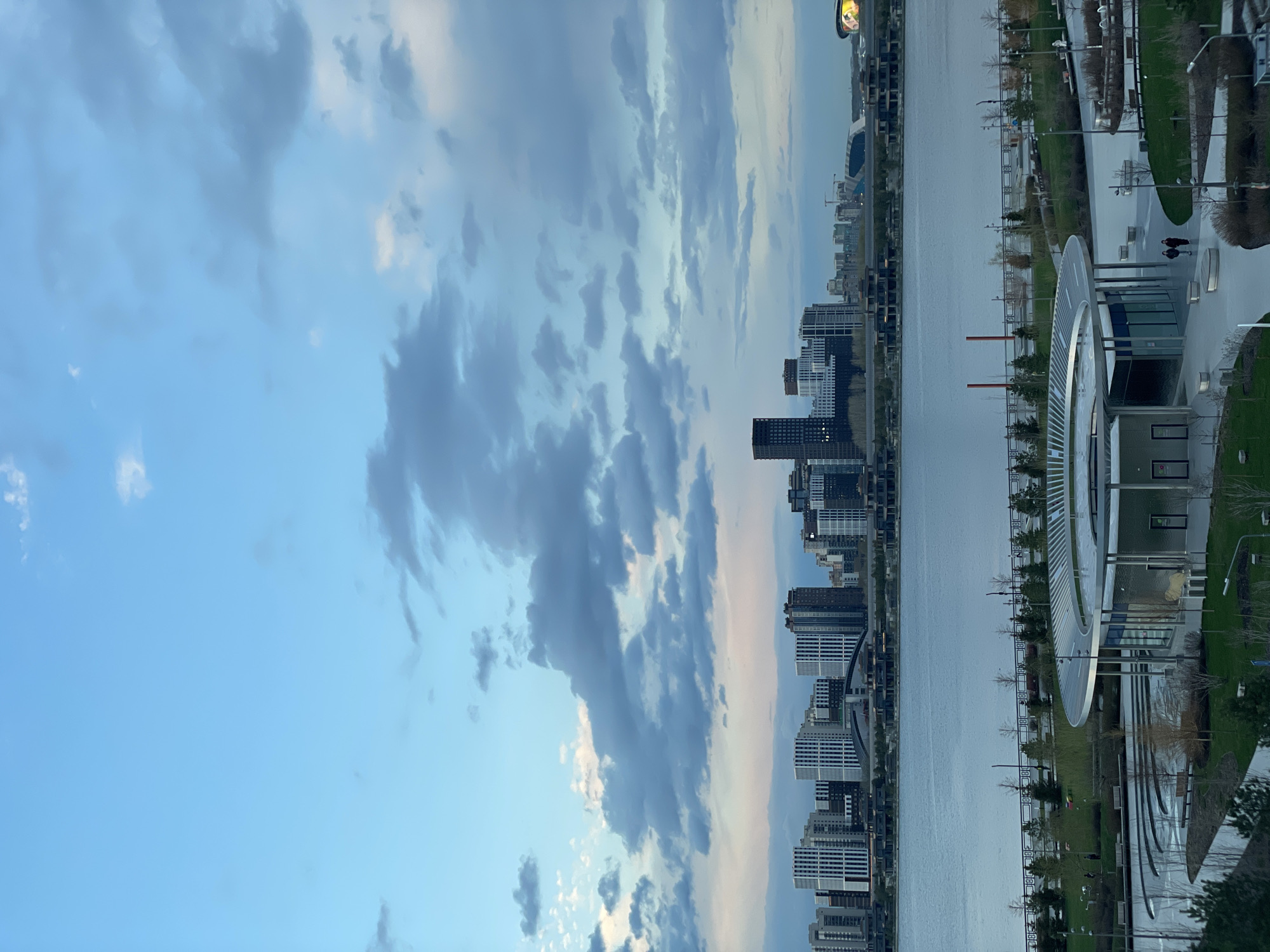 A panoramic shot captures a modern cityscape along a wide river or canal under a cloudy sky. The upper portion of the image is dominated by a blue sky filled with varied cloud formations, ranging from dark grey to soft white cumulus clouds.
A wide, calm body of water, likely a river, horizontally bisects the scene, reflecting the sky's muted light. On the far side of the river (left), a dense skyline of numerous tall buildings and skyscrapers, featuring diverse architectural designs in shades of grey and white, forms a busy urban backdrop.
On the near side of the river (right), a spacious waterfront park or promenade unfolds. It includes wide, paved pathways, neatly manicured green lawns, and evenly spaced trees and streetlights. Two individuals are seen walking casually on one of the pathways in the mid-right area. A striking modern architectural structure with a large, curved, silver-toned arch or roof is prominent on the right, appearing to house a building or pavilion. Further in the distance on the far right, a large, circular or oval structure, possibly part of a stadium or a public venue, is visible with a brightly lit, yet unreadable, sign at its peak.
The soft, diffused lighting suggests it is late afternoon or early evening. The scene portrays a tranquil urban environment. No discernible text is visible within the image beyond the blurry illuminated sign.