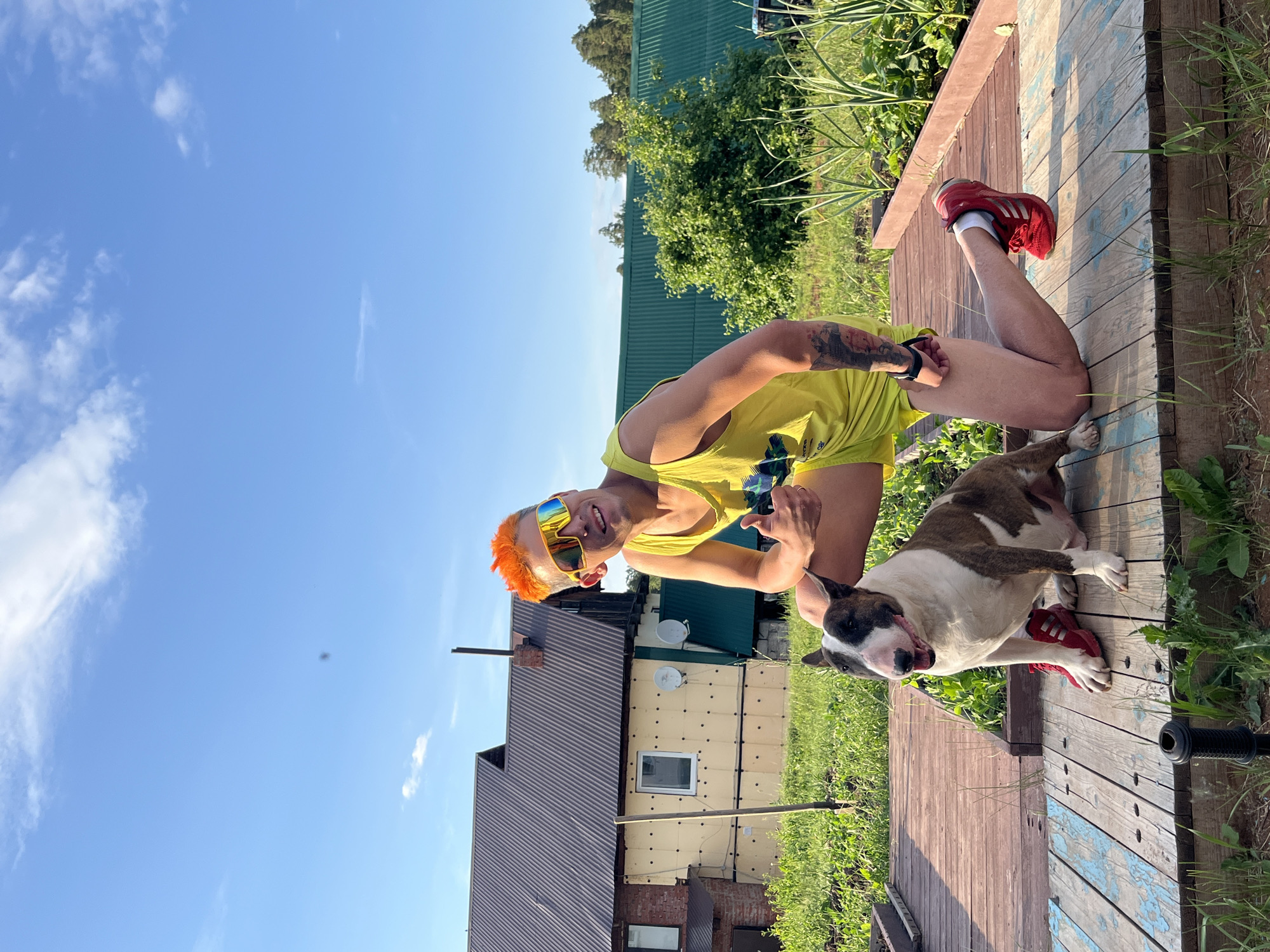 In a backyard setting in the city of Chaikovsky, Russia, a man with bright orange hair and yellow sunglasses kneels on a wooden deck. He is wearing a yellow tank top and shorts, and gives a thumbs-up gesture. Beside him, a bull terrier dog sits on the deck, wearing red sneakers. In the background, a house with a dark metal roof and light-colored siding is visible, along with greenery and a bright blue sky with scattered clouds.