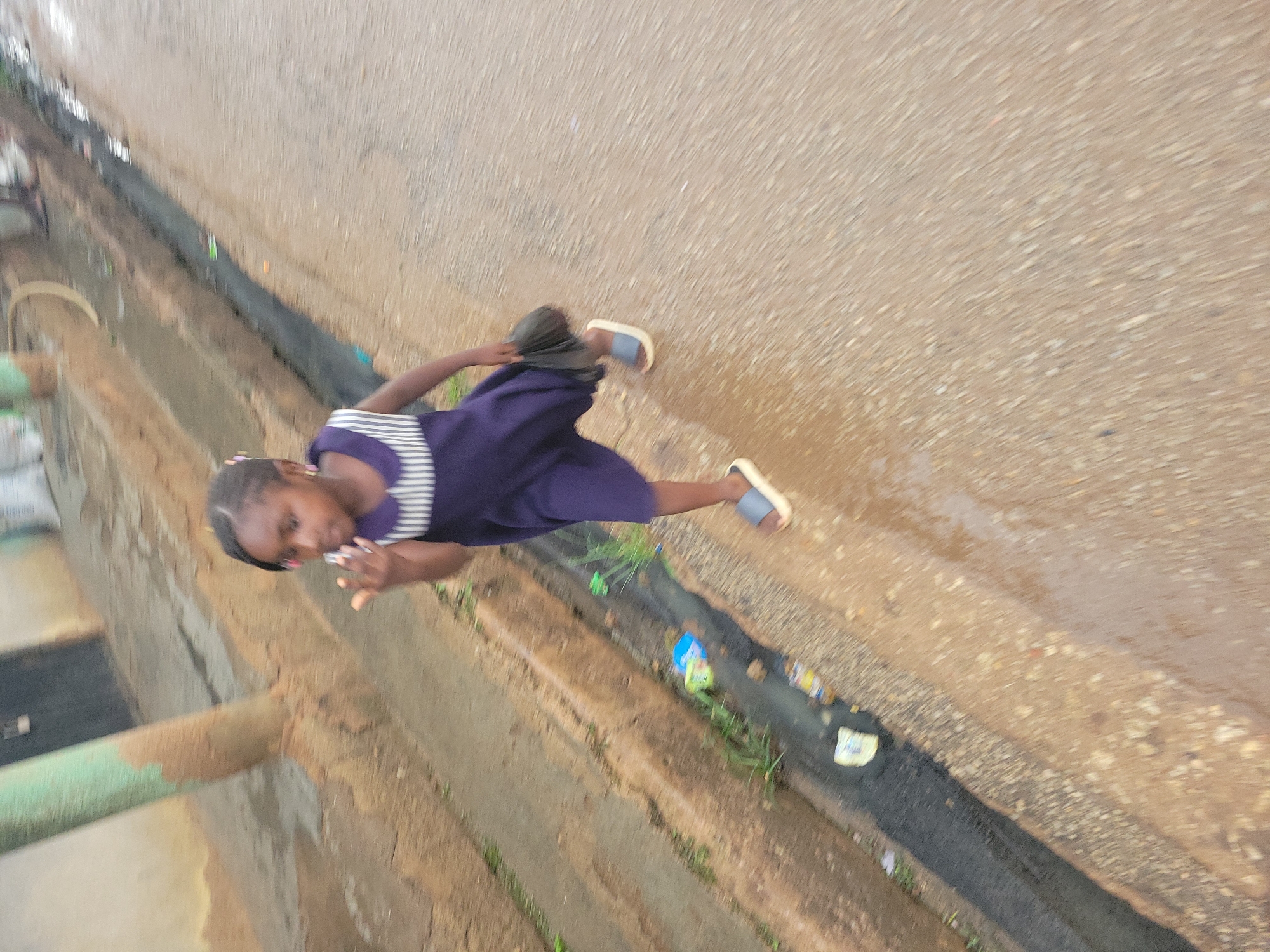 The image shows a young girl walking on a wet street in Jos, Nigeria. She is wearing a dark purple dress with a white striped collar and white sandals. She is carrying a dark object in her right hand. The street appears to be made of asphalt and is wet, with puddles visible. There is a drainage ditch on the left side of the street, with some trash and vegetation in it. A light-colored wall and a concrete structure are visible in the background. The image is slightly blurred, suggesting movement.