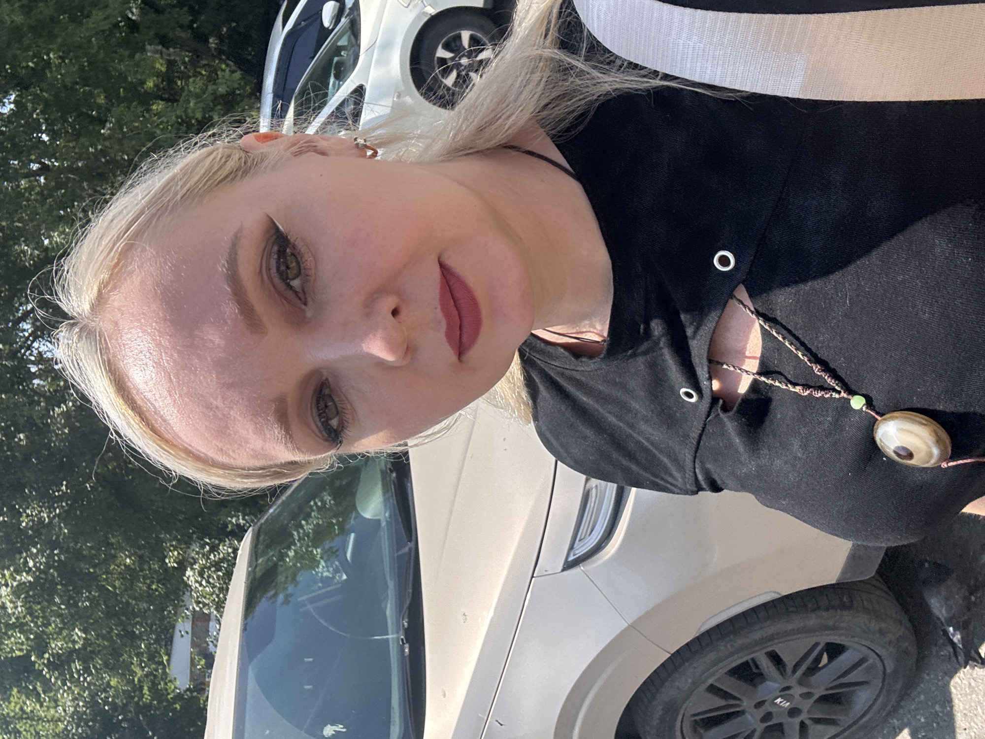 A woman with blonde hair is taking a selfie outdoors. She is wearing a black t-shirt with a cut-out design and a necklace with a large pendant. A light-colored SUV is parked behind her, and another white car is visible further in the background. Trees and foliage suggest a location with natural surroundings, possibly a park or residential area. The sun is shining, casting shadows. The scene appears to be in Komsomolsk-on-Amur, Russia.