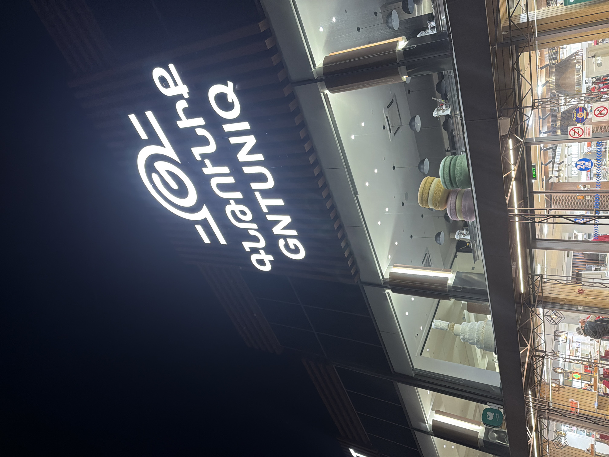 The image shows a storefront at night, with a brightly lit sign reading "GNTUNIO @nature" vertically displayed. Inside the store, there are hanging decorations resembling macarons and chandeliers, and shelves are visible with various merchandise. People are inside the store, browsing the items. The location is most likely in Aparan, Armenia, based on the sign's language.