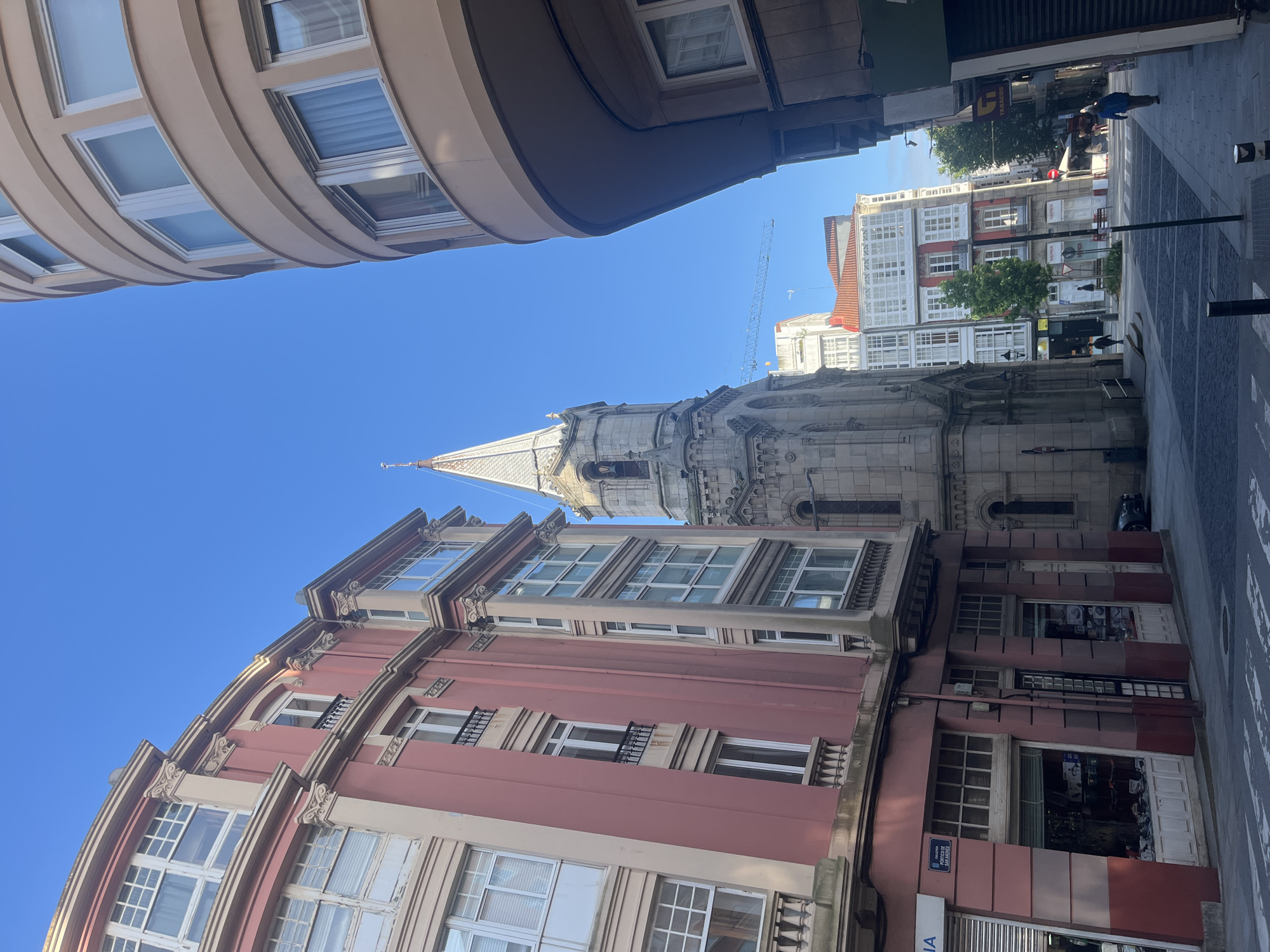A street view in A Coruña, Spain, presents a perspective looking upwards between buildings towards a church spire. The architecture on the left features a pink facade with rows of windows. Above and to the left, a curved building overhangs the street. In the distance, other buildings with white facades are visible. A crane can be seen among the buildings. The sky is clear blue. A section of the sidewalk and street is visible on the right.