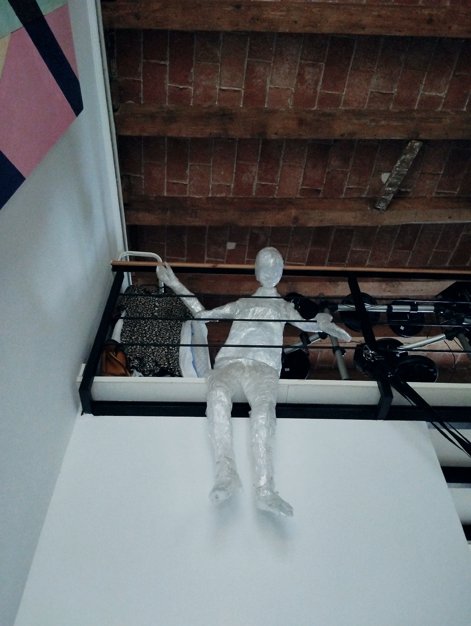 The image captures an indoor scene with a visible mezzanine or elevated platform. A mannequin, seemingly wrapped in translucent plastic, is positioned on the edge of this platform, with its legs dangling. The mannequin is in a seated or slumped posture. Below the platform, on the surface of what appears to be a wall, is a abstract geometric artwork featuring pink and dark blue shapes.
The background above the platform reveals a ceiling constructed of exposed wooden beams and terracotta tiles, characteristic of traditional Spanish architecture. The mezzanine itself features a black metal railing and a wooden handrail. Various items are visible on the platform behind the mannequin, including clothing (a patterned dress and a white garment), a brown handbag, and some electronic equipment with cables. The overall impression is of an interior space with a rustic ceiling and a modern mezzanine, possibly an apartment, studio, or gallery in Barcelona, Spain. The lighting suggests an indoor setting, possibly during the day given the ambient light.