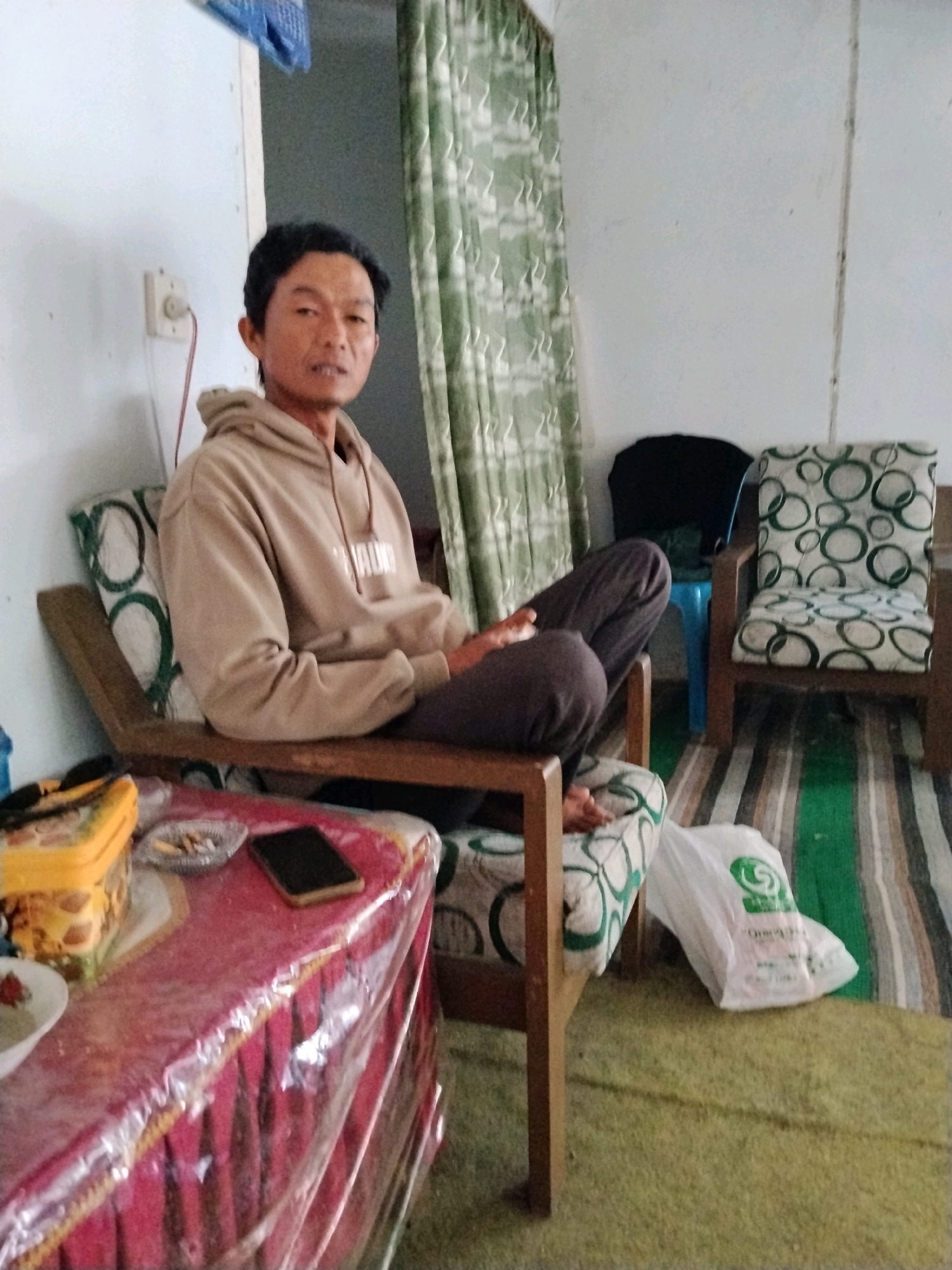 In an indoor residential setting, likely a living room, a middle-aged East Asian male is seated on a wooden chair with a patterned cushion. He is wearing a light brown hooded sweatshirt and dark pants. The room features a white wall with an electrical outlet and a light green patterned curtain. To the right, another wooden chair with a similar cushion is visible, along with a blue plastic stool and a black object, possibly a bag. A carpeted floor with striped runners is present. In the foreground, a table is covered with a red cloth and a clear plastic sheet, displaying a yellow container, a small ashtray with cigarette butts, and a dark-colored smartphone. A white plastic bag with a green logo lies on the floor near the chair. The scene appears to be in Panggungan, Indonesia, based on the provided context.