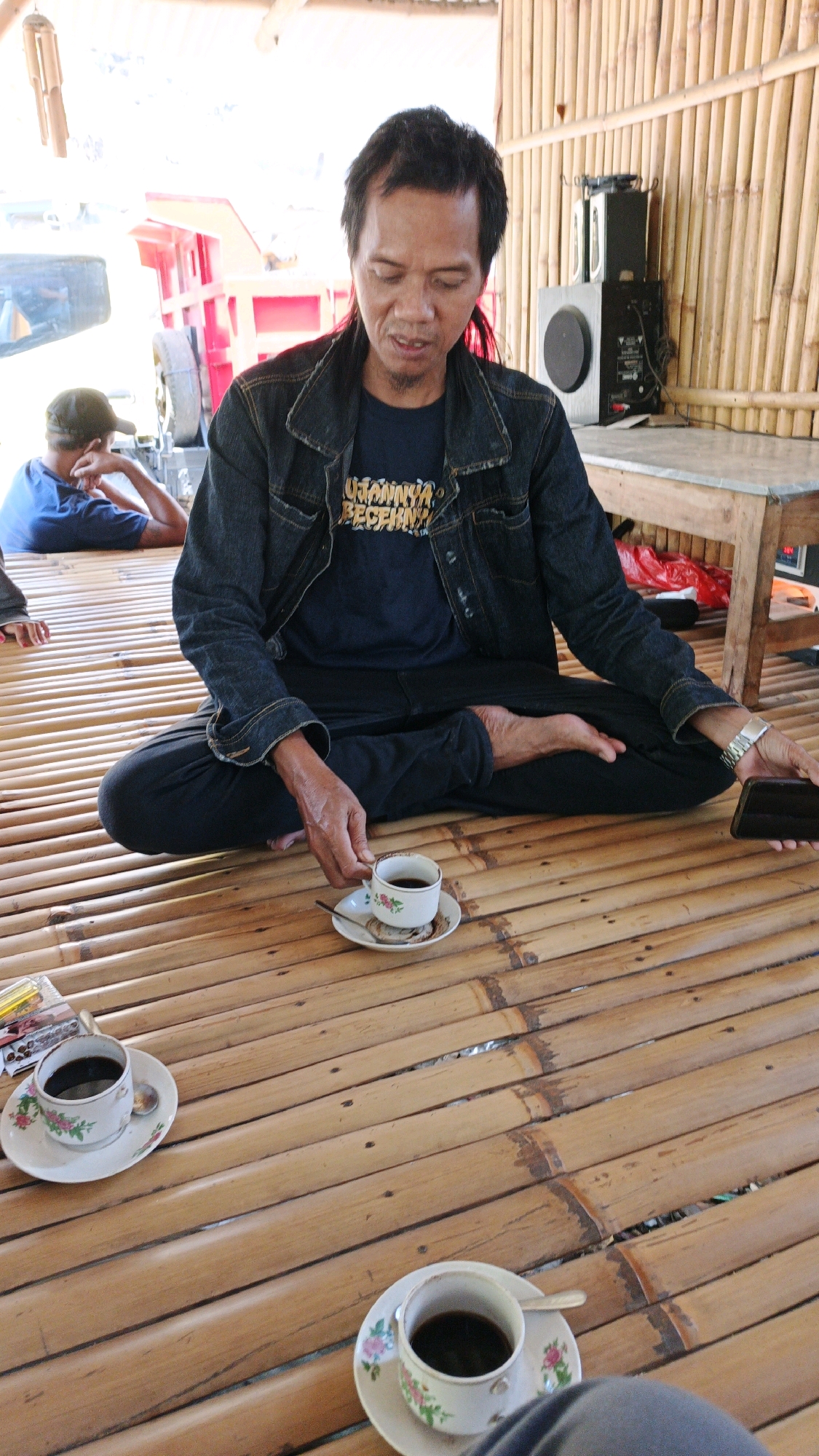 A man is sitting cross-legged on a bamboo floor in Sanggrahan, Indonesia, with a cup of coffee on a saucer in front of him. He is wearing a denim jacket and a dark blue t-shirt. He is holding a phone in his right hand and a cup of coffee in his left hand. There are two other cups of coffee on the floor, one to his left and one in the foreground. A young man in a blue t-shirt and a cap is sitting in the background, facing away from the camera. The environment is rustic, with bamboo walls and a wooden table.