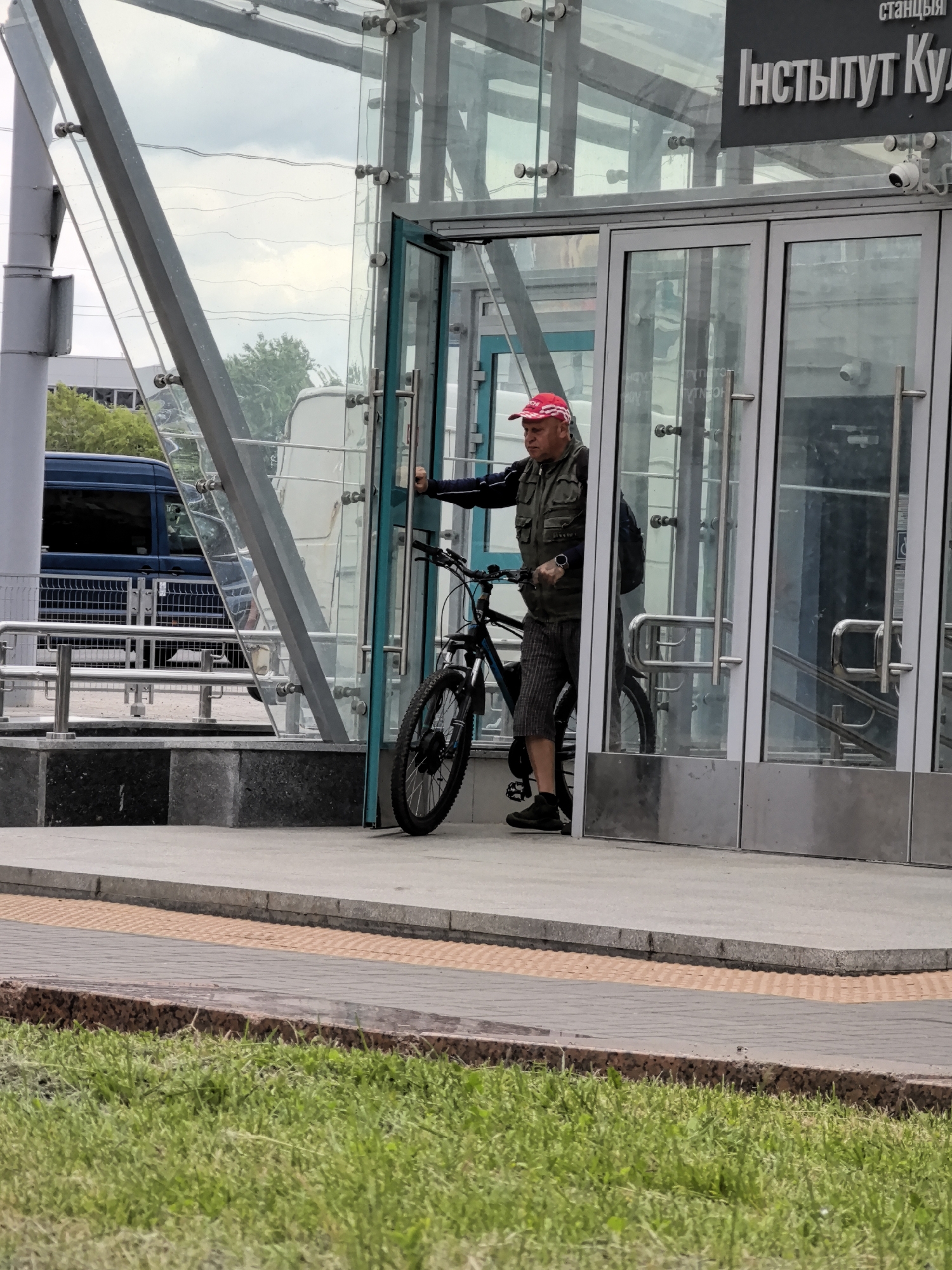 A man wearing a red and white striped cap, a green vest over a dark long-sleeved shirt, and plaid shorts is pushing a bicycle next to the entrance of a building with glass doors. He is standing on a concrete walkway in front of the building, which has a sign above the doors with Cyrillic text. A dark blue van is visible through the glass of the building. The scene appears to be outdoors, with some grass in the foreground.