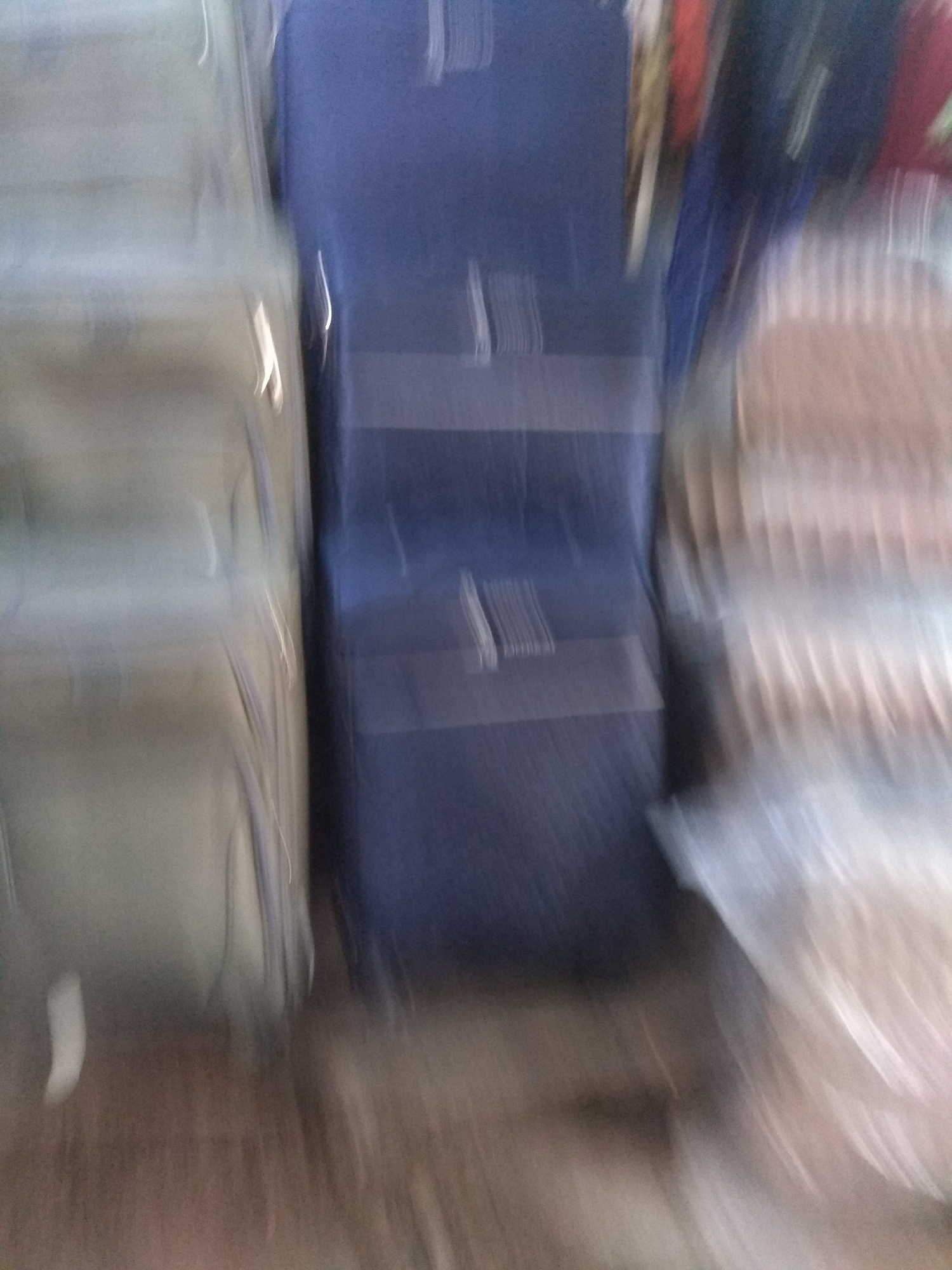This is a blurry, action-shot photograph of several suitcases. Two large, light-colored suitcases are on the left, and a dark blue suitcase is on the right, positioned in front of what appear to be stacks of other luggage. The suitcases are upright and appear to be made of fabric or a soft material. The overall impression is of a collection of travel bags, possibly in a storage area, shop, or transport hub. The motion blur suggests movement, either of the camera or the subjects themselves. The background is indistinct, but hints of other items and colors are visible, suggesting a busy environment. There is no visible text in the image. The time of day and weather are not discernible. The emotion conveyed is difficult to ascertain due to the blurriness, but the image might suggest a sense of travel or transition.