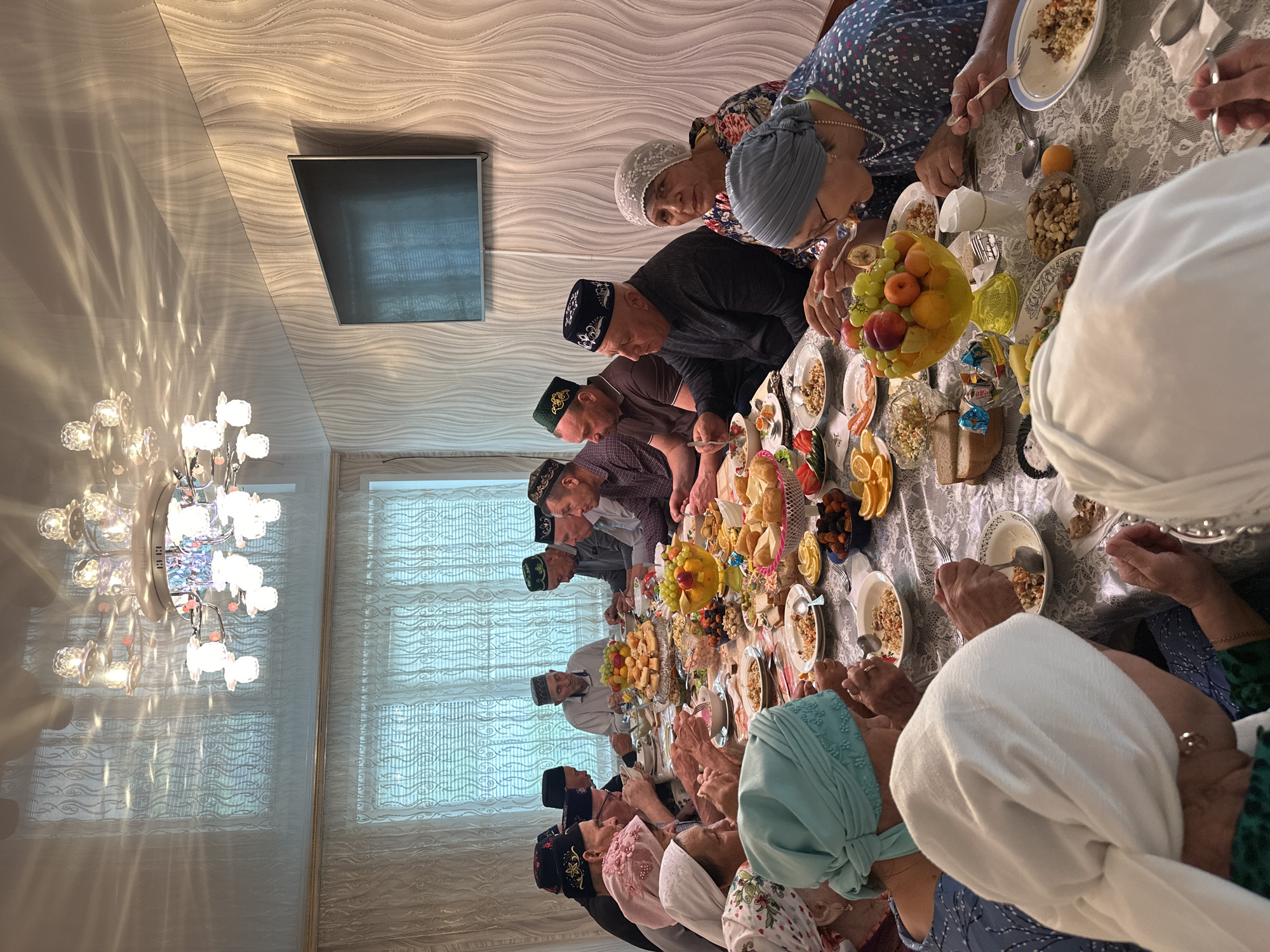 A group of people, both men and women, are gathered around a long table laden with food. The men are wearing traditional skullcaps, while many of the women wear headscarves. The table is covered with a lace tablecloth and holds a variety of dishes including fruits, nuts, salads, and bread. Some people are eating, using spoons to serve themselves from the dishes. The setting appears to be indoors, likely a dining room, based on the chandelier overhead and the window with sheer curtains. A television hangs on the wall. The location is Коноваловка, Russia.