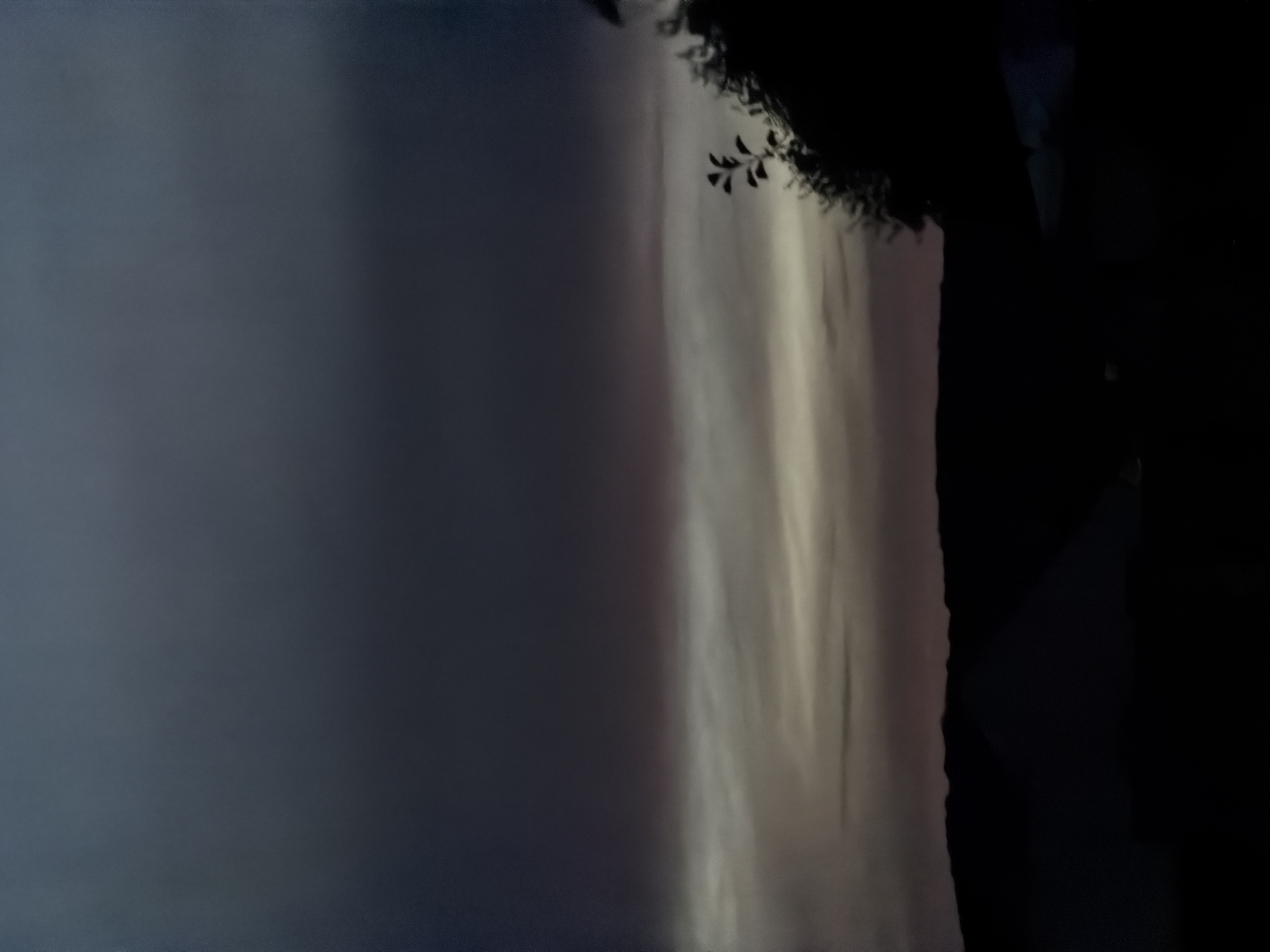 This is a low-light photograph of a natural landscape at dusk. The sky occupies the majority of the frame, transitioning from a dark, deep indigo at the top to lighter shades of gray and pale pink near the horizon. Thin, wispy clouds are visible in the middle ground, catching the last vestiges of daylight. Below the sky, a dark silhouette of rolling hills forms the distant background. In the foreground, the silhouette of trees and foliage is visible on the right side of the image, with a few indistinct shapes that could be structures or tents on the left. The overall atmosphere is somber and still. The image was taken in Junglinster, Luxembourg.