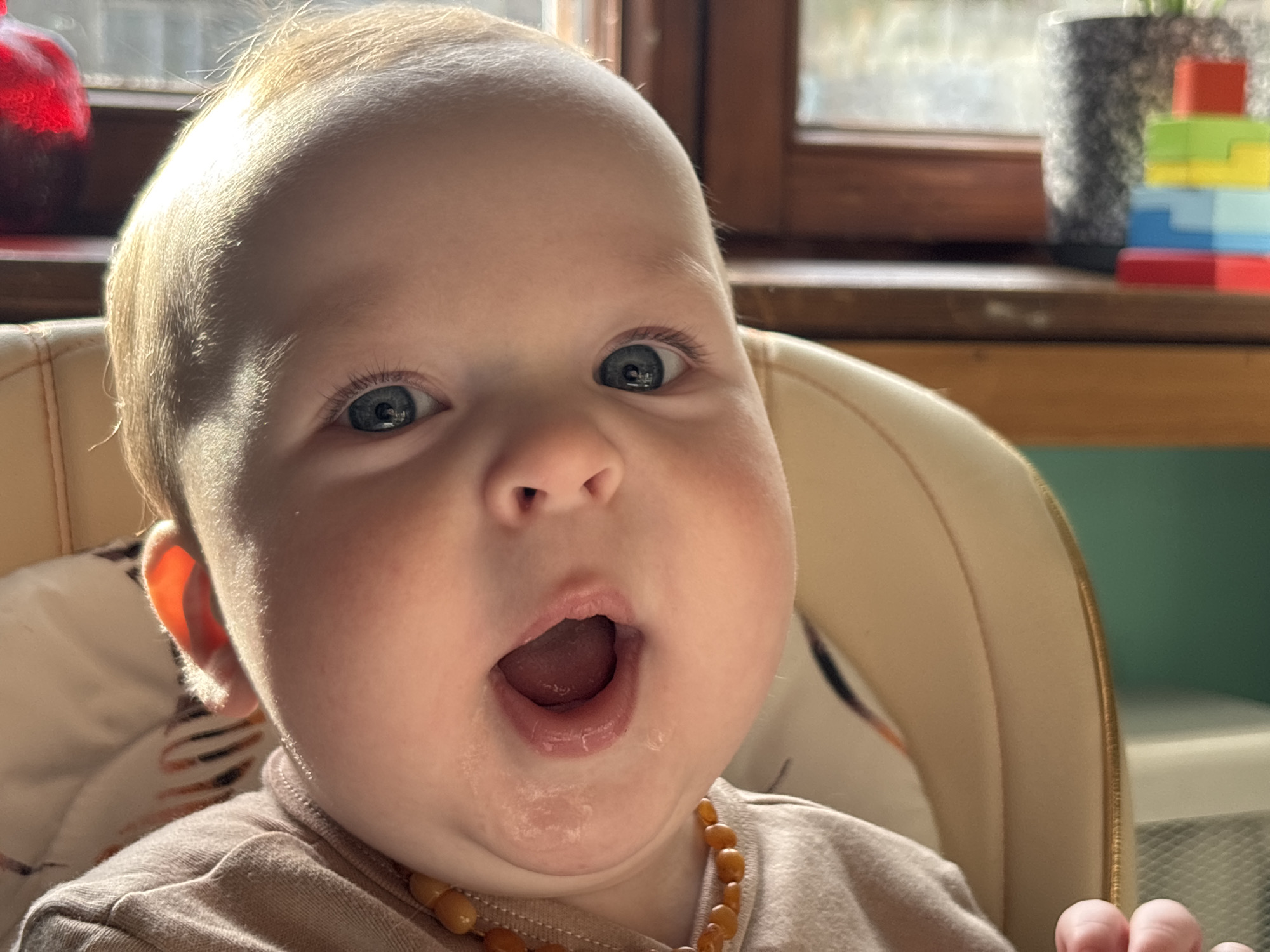 A close-up, brightly lit photograph shows a baby sitting in a high chair, looking directly at the camera. The baby has fair skin, light-colored hair, and wide blue eyes. Its mouth is open in an expression of surprise or awe, with visible drool on the chin. The baby wears a light brown long-sleeved top and a necklace made of amber-colored beads.
Strong natural light, likely from a window, illuminates the baby's face, hair, and translucent ear, creating highlights. In the background, to the right, a wooden window frame is visible. On a surface beneath the window, a dark gray textured pot containing a plant and a colorful stacking toy with red, green, yellow, and blue blocks are partially in view. To the left, a blurry red object can be seen. The high chair is a light beige or cream color. The setting appears to be an indoor home environment during the daytime in Марковский, Russia. There is no visible text in the image.