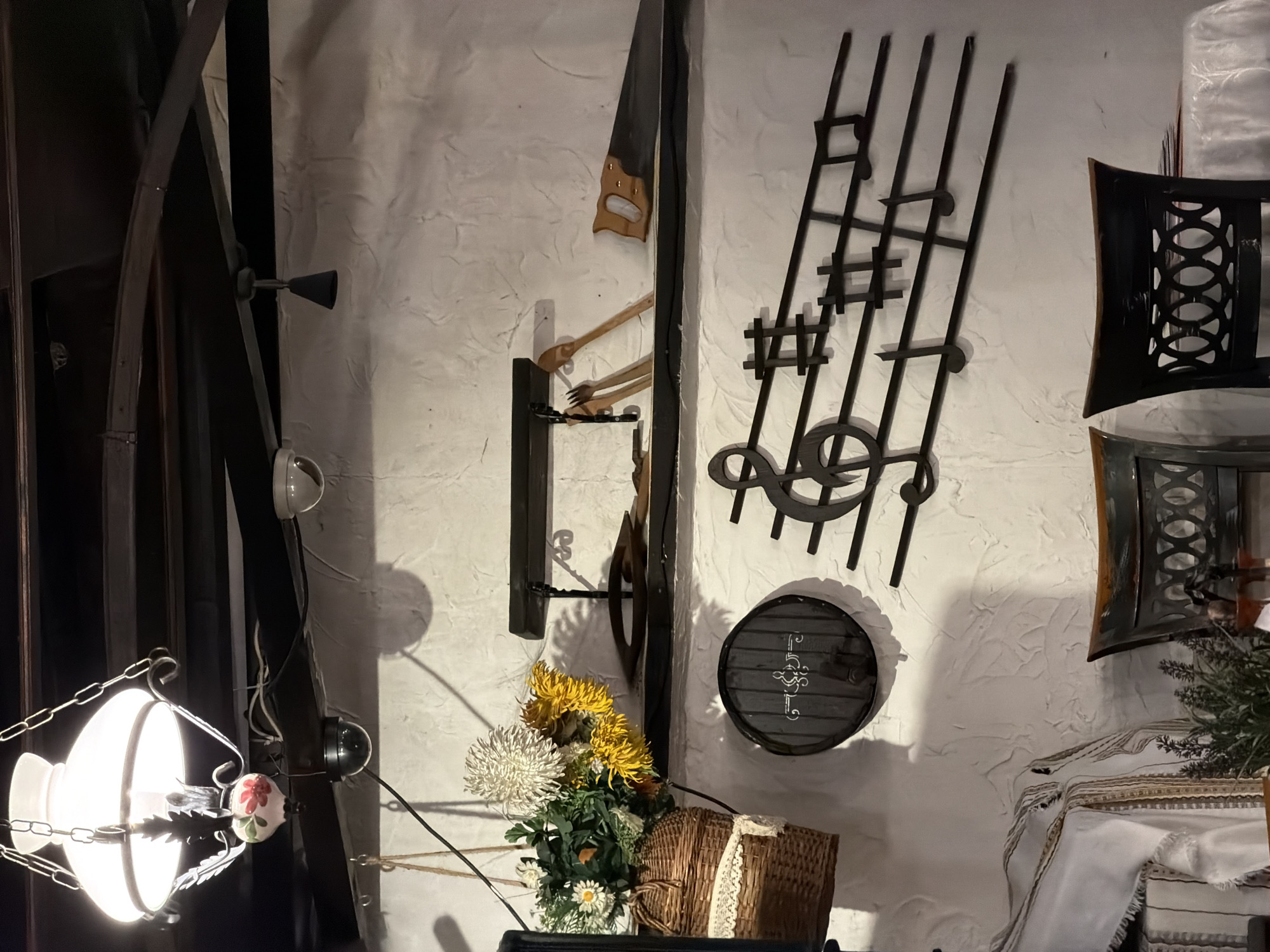 This is an interior shot depicting a decorative wall arrangement. The wall, with its textured, white surface, features several items. On the left, a complex dark metal framework is visible, incorporating a hanging lamp with a white, bulbous shade. To the right of this, a saw and utensil display hang on the wall. Further right, there's a dark metal music note design. Below the music notes, a round, dark metal object with lighter markings hangs. In the foreground, a basket of flowers is visible. To the right, there is a chair back of decorative metal and fabric.
The location is Varna, Bulgaria.
