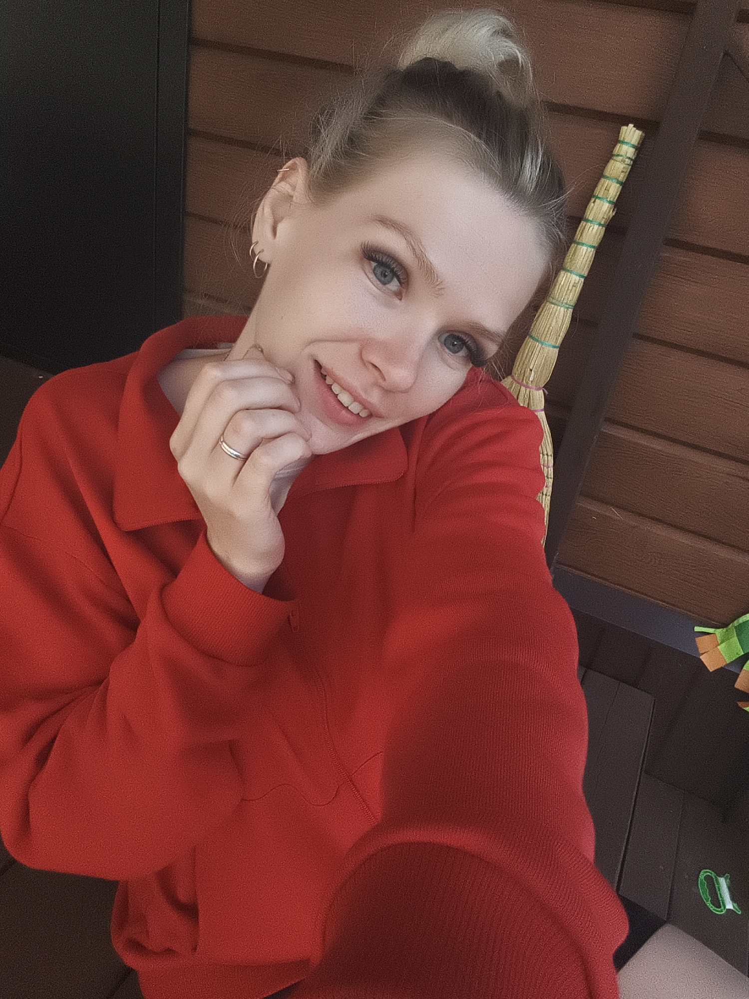 A young woman with blonde hair pulled into a bun smiles at the camera, resting her hand on her cheek. She wears a red hooded sweatshirt. Behind her, a wooden structure with horizontal planks is visible, and a broom leans against it. The environment appears to be outdoors, possibly a porch or balcony. The overall lighting suggests a daytime setting.