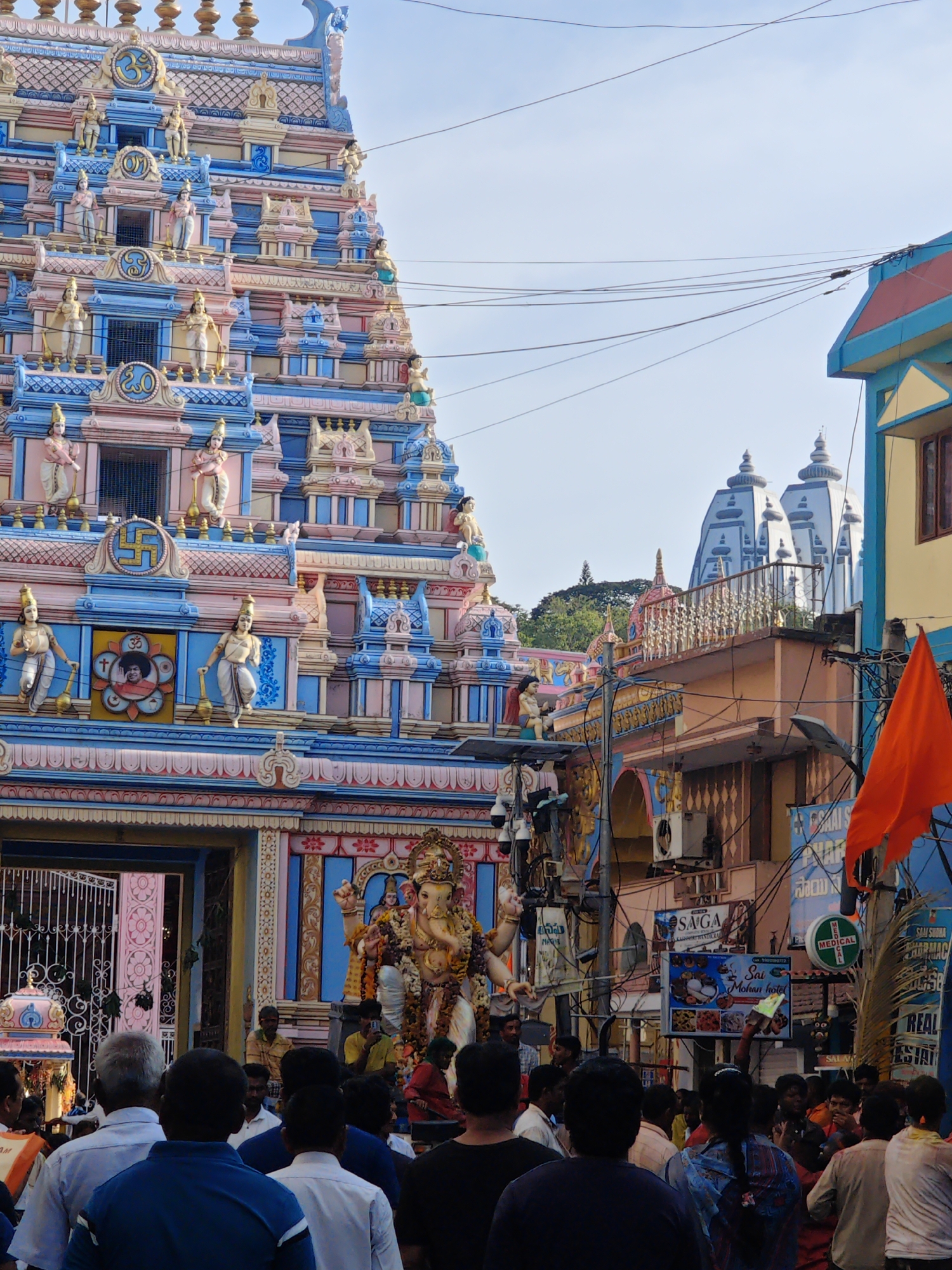 The image depicts a street scene in Puttaparthi, India, dominated by the colorful and ornate architecture of a Hindu temple. The temple's facade is intricately decorated with numerous statues of deities and figures, painted in vibrant pinks, blues, and gold. Above the entrance, a large statue of Ganesha, the elephant-headed deity, is visible, adorned with garlands. A crowd of people, dressed in casual attire, are gathered in front of the temple, some appearing to be devotees or tourists. The street is also lined with shops and buildings, with electrical wires crisscrossing overhead, indicative of an urban environment. An orange flag is prominently displayed on the right side of the image. The overall atmosphere suggests a lively and bustling religious or cultural event taking place in a typical Indian town.