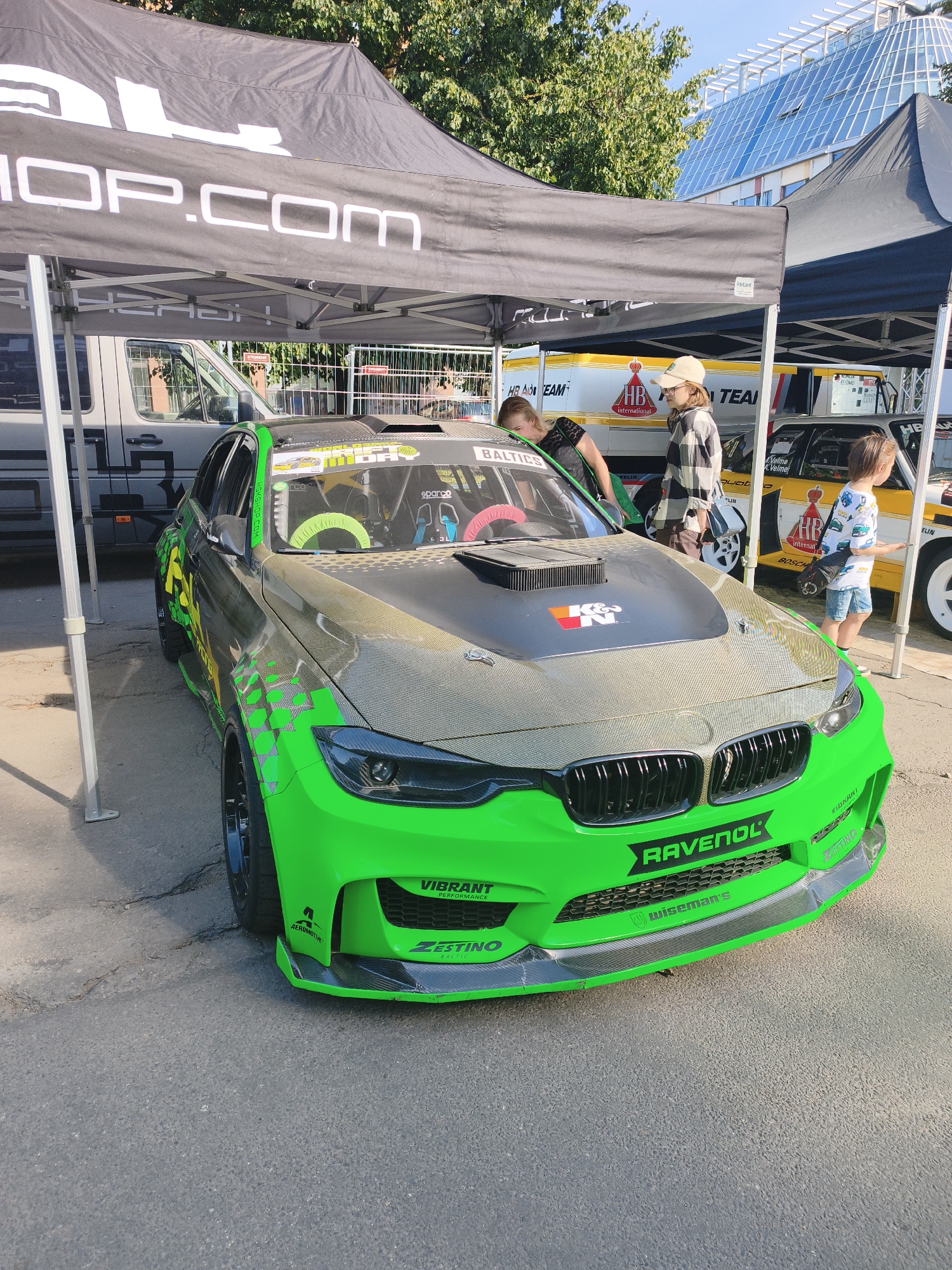 The image depicts a modified green racing car parked outdoors, possibly at a motorsport event or car show. The car, which appears to be a BMW, is adorned with sponsor logos such as "RAVENOL", "VIBRANT PERFORMANCE", and "ZESTINO". It has a carbon fiber hood and extensive aerodynamic modifications. Several people are visible around the car, including individuals interacting with the vehicle and others in the background. A tent with the text "SHOP.COM" is set up over part of the scene. In the background, a modern glass-building is visible, suggesting an urban setting. The presence of a yellow racing car with "HB TEAM" branding further indicates a motorsport-related gathering.
The specific location of this image in Riga, Latvia, is the Riga Motor Museum (Rīgas Motormuzejs), a popular venue for car enthusiasts and events. The museum is known for its extensive collection of vintage and modern vehicles, and it frequently hosts events that would draw such a scene.