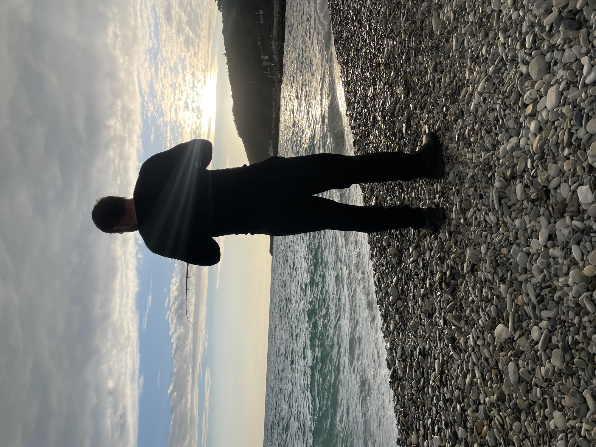 The image depicts a person standing on a pebble beach in Olginka, Russia. The person is silhouetted against the setting sun, which is partially obscured by clouds in the sky. The waves are visible along the shoreline, and a coastal building can be seen in the background. The individual appears to be holding a fishing rod. The ground is covered in pebbles and small rocks, while the sky displays clouds, with the sun setting in the background, its rays reflecting on the water.