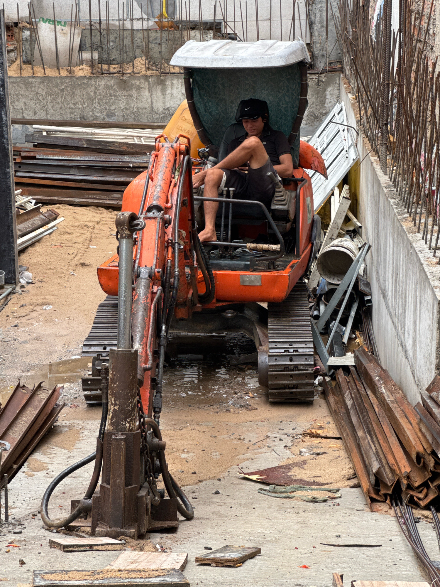 The image depicts a construction site in Nha Trang, Vietnam, with an excavator as the central subject. A man wearing a black t-shirt and shorts sits in the driver's seat of the orange excavator, appearing to be taking a break. The excavator is positioned on a sandy and muddy ground, surrounded by construction materials like rebar, metal beams, and scaffolding. The background shows unfinished concrete walls and structures, indicative of ongoing construction. The overall scene is one of an active, albeit momentarily paused, construction project in an urban setting.